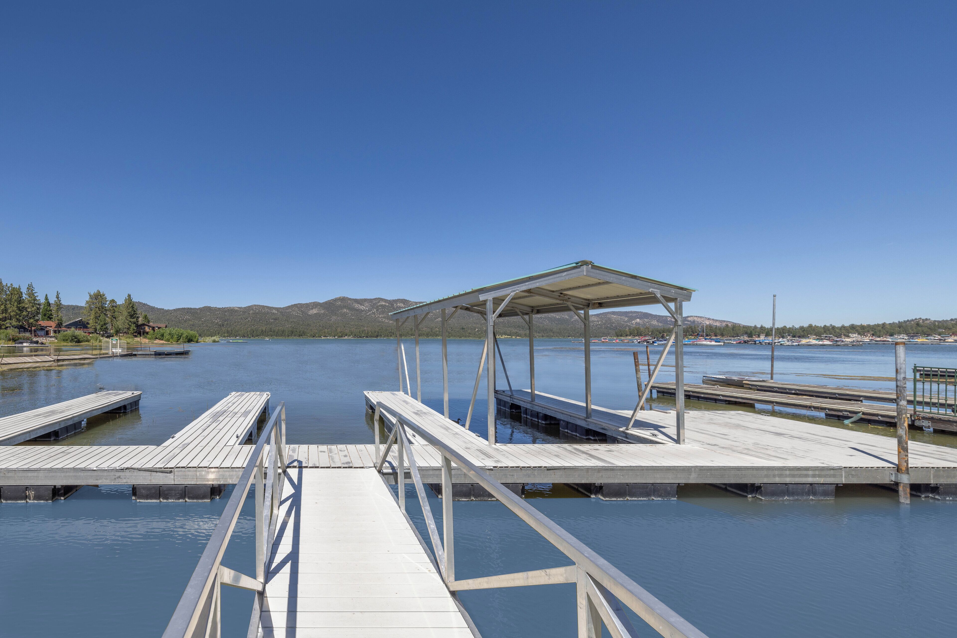 A wooden pier leading to a covered seating area by a calm lake with mountains in the background.