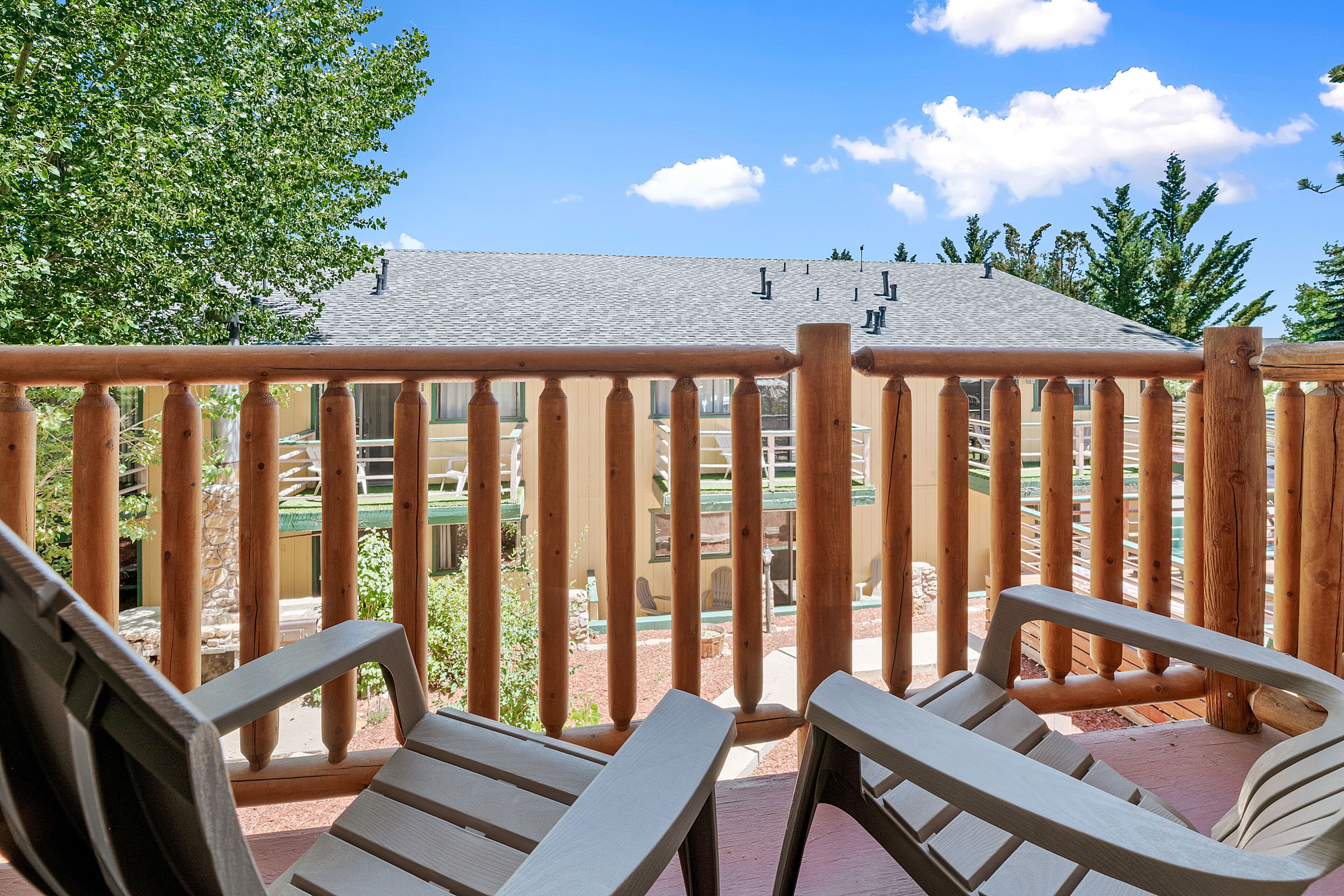 A wooden deck with two Adirondack chairs, a stone wall, and a view of a building with a grey roof.