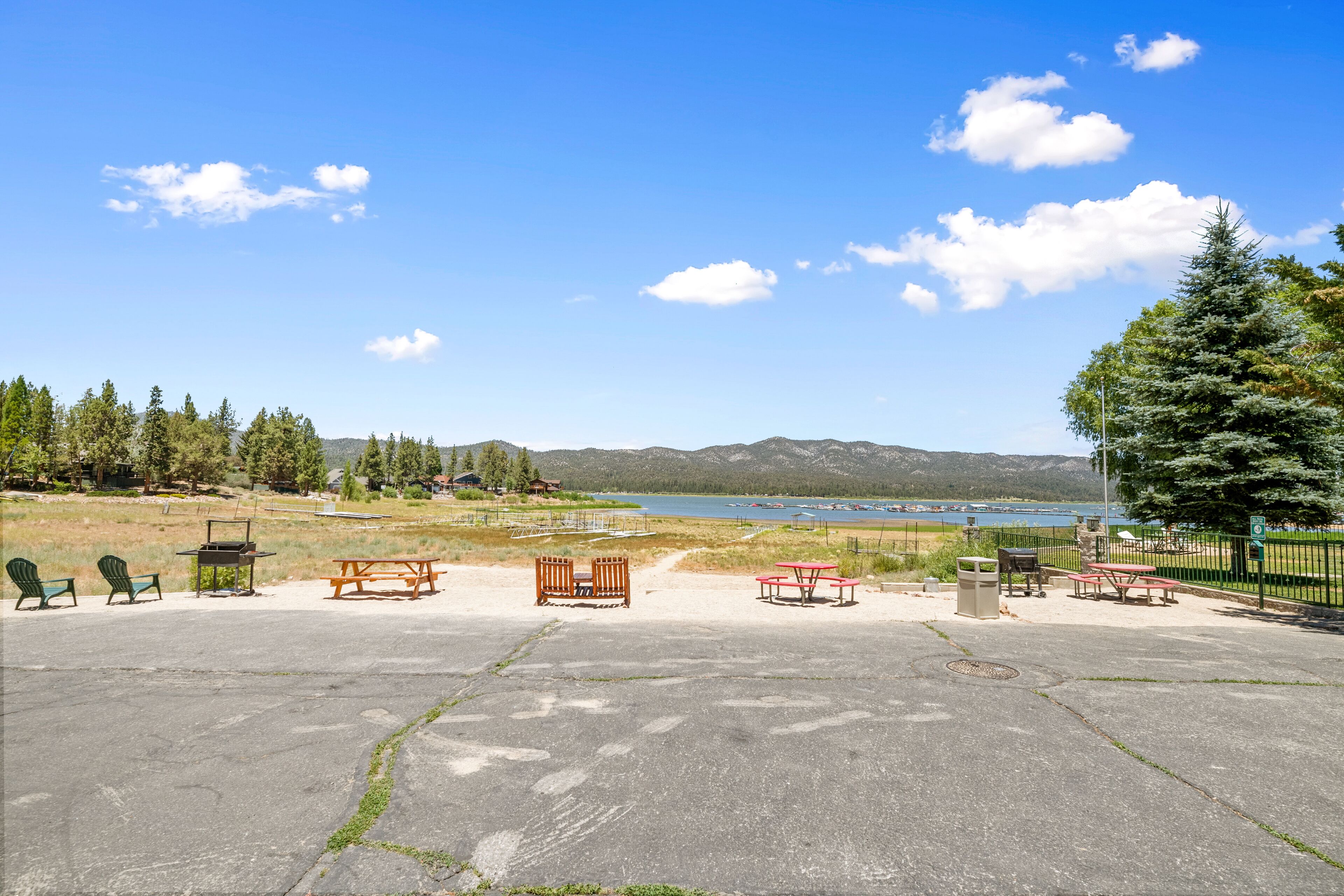 A concrete parking area with picnic tables and chairs, a fenced area with trees, and a view of a lake and mountains in the background.