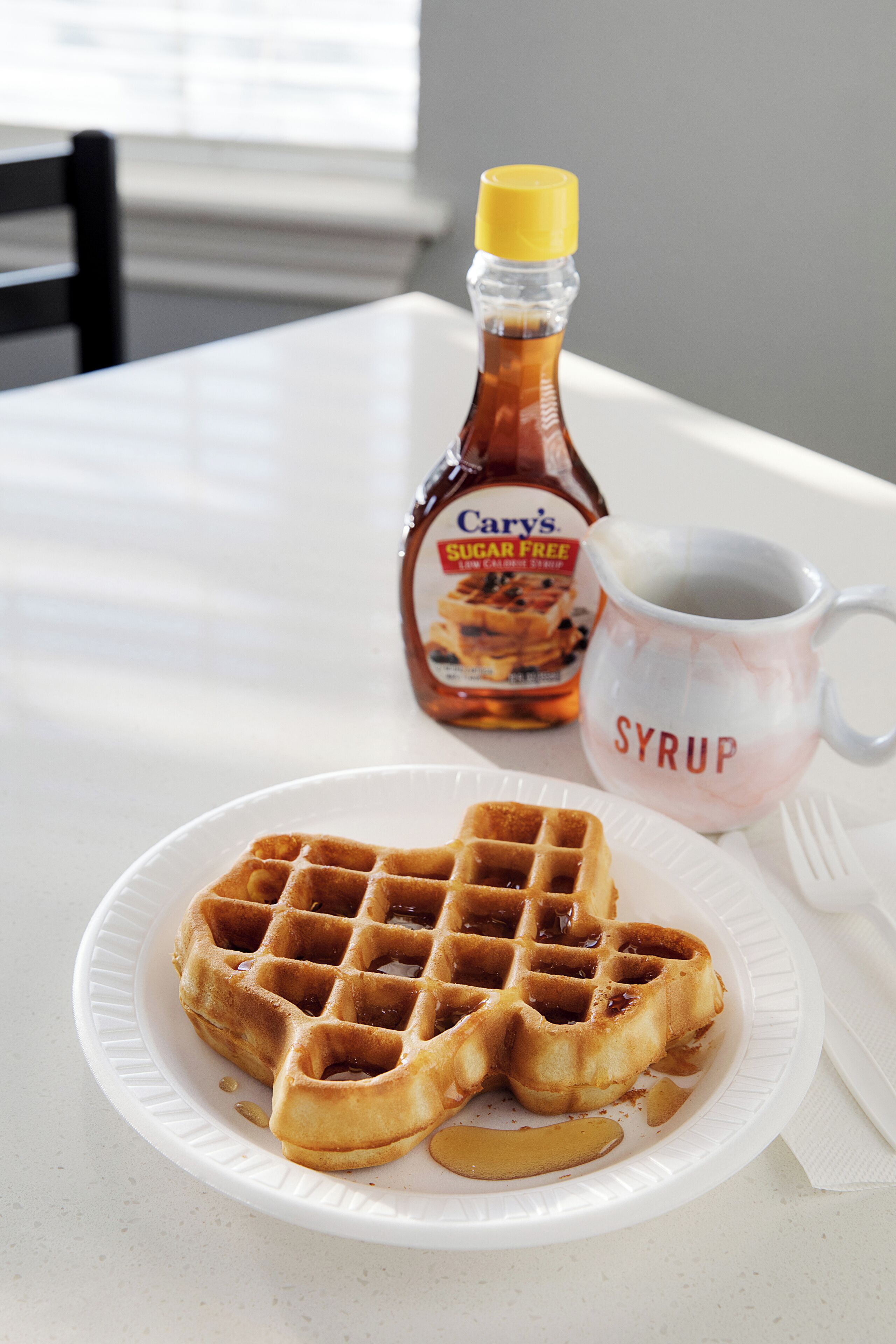 Un plato con waffles y jarabe, una botella de jarabe y una taza con la etiqueta 'SYRUP' sobre una mesa.