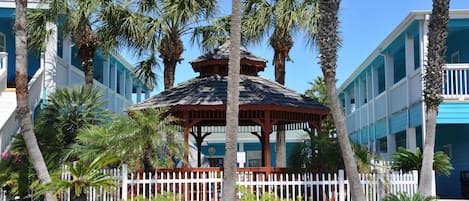 A gazebo surrounded by a white picket fence, palm trees, and a building with blue and white exterior walls.