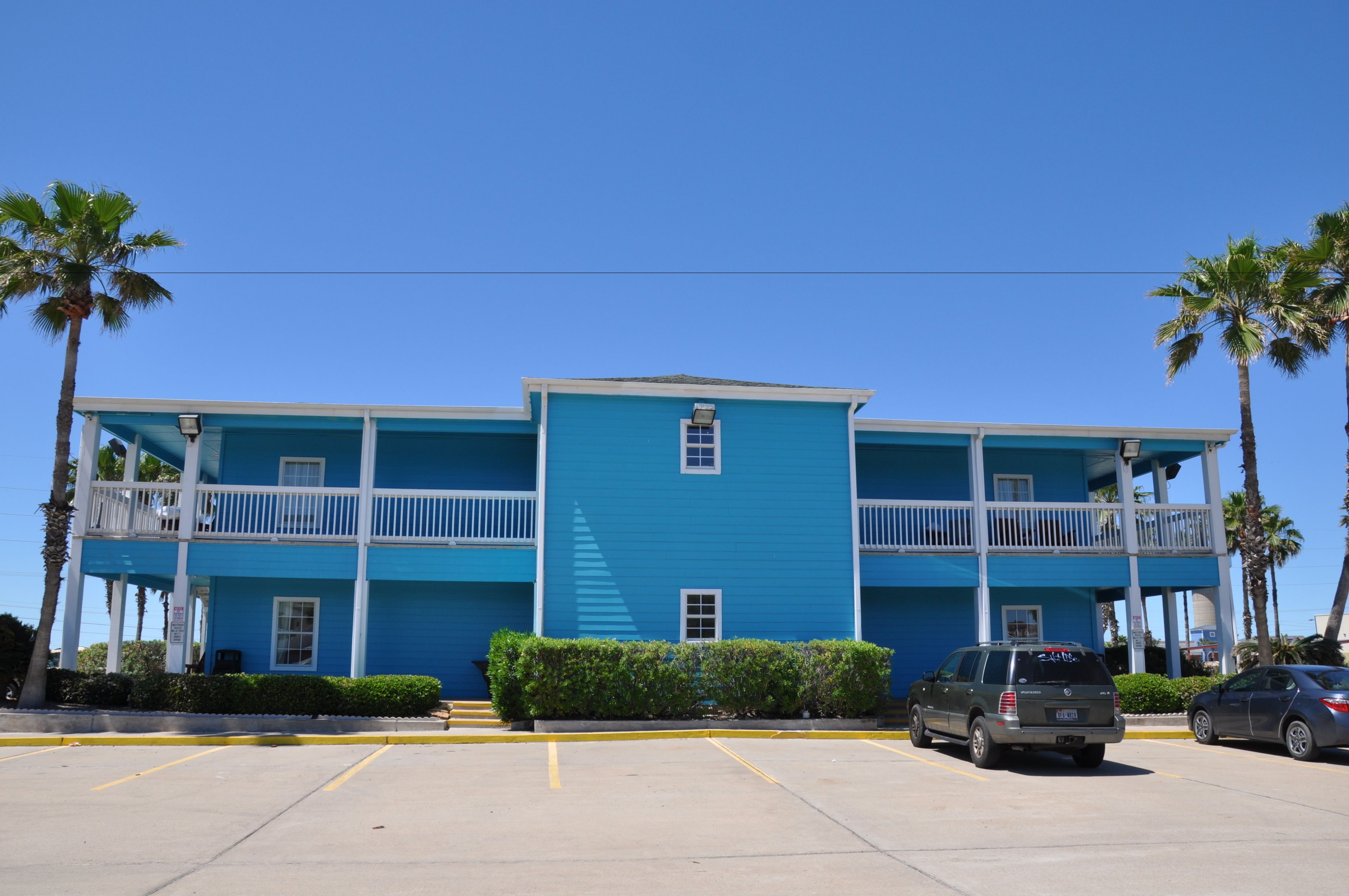 A two-story building with a blue exterior and white balconies. There are palm trees and a parking lot in front.
