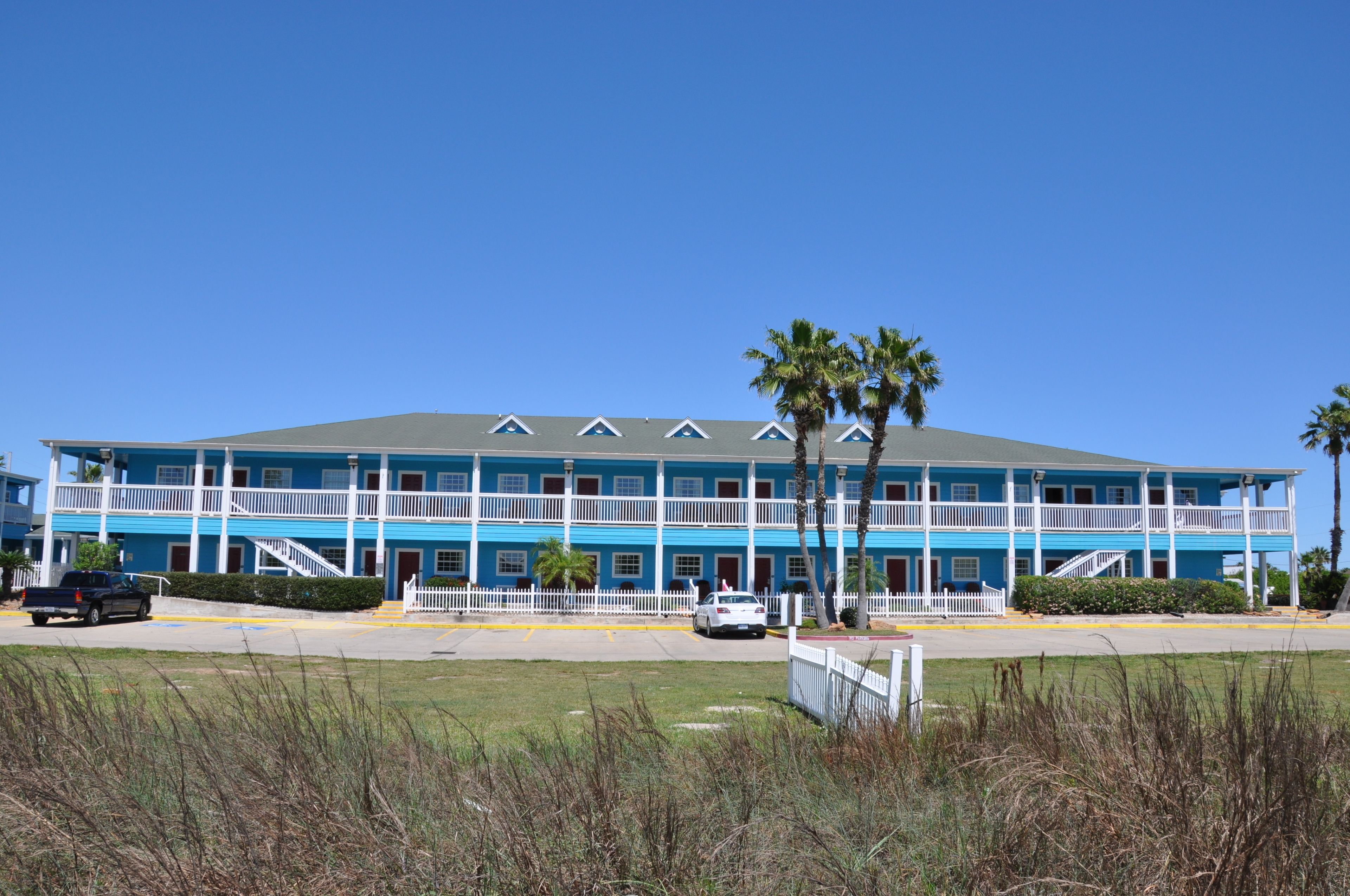 A two-story motel with a blue exterior and white railings, featuring a central staircase, palm trees, and a parking lot.