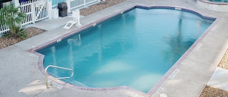 A swimming pool with lounge chairs and a gazebo, surrounded by a fenced area with a building in the background.