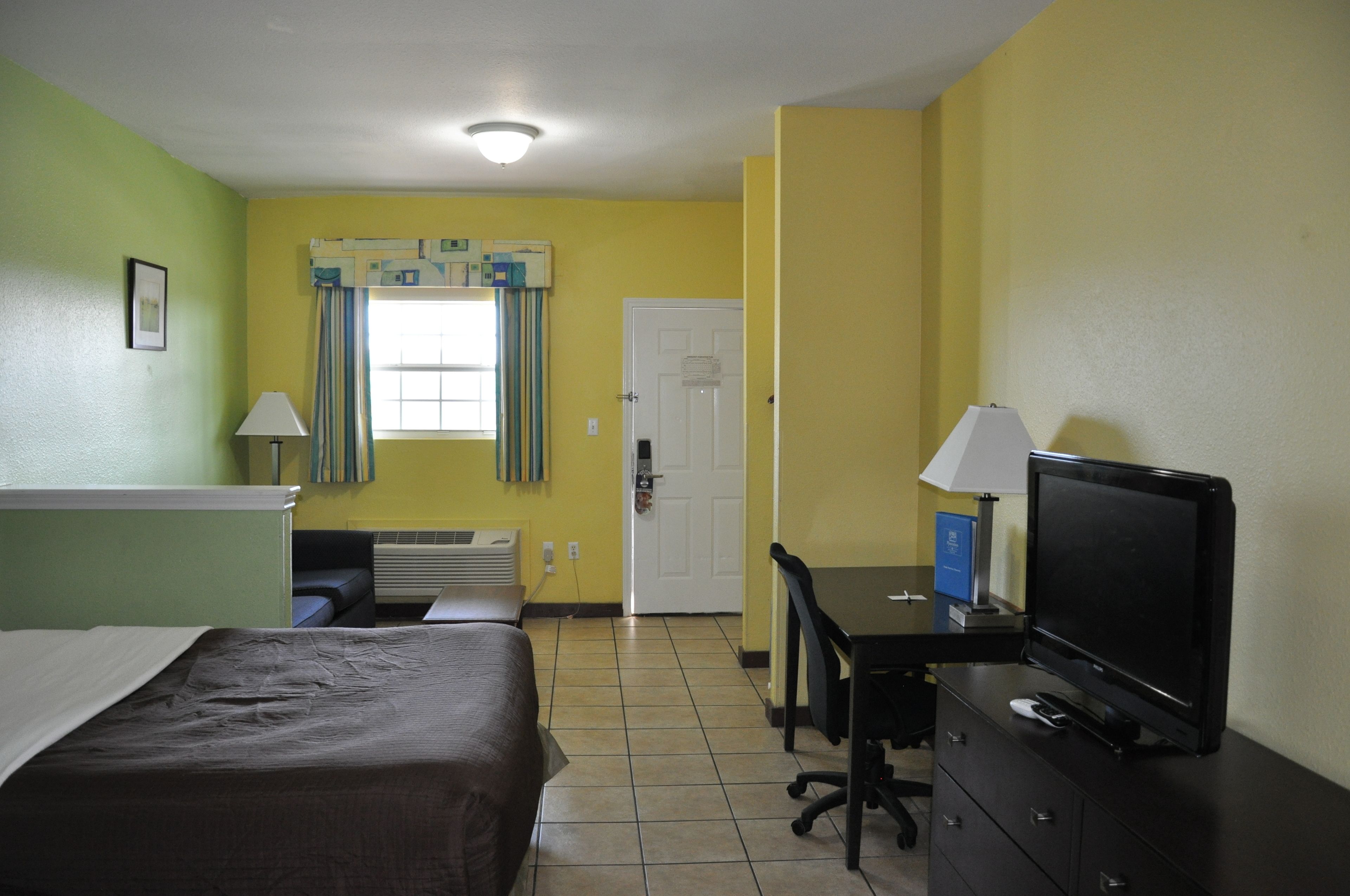 A hotel room with a bed, a desk with a computer and TV, a window with curtains, and a door.