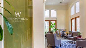 A lobby with a glass wall displaying the logo "WAGON WHEEL LOUNGE", a seating area with chairs and a table, and large windows allowing natural light. - Church Street Inn Historic Charleston, Ascend Collection Hotel (Charleston)