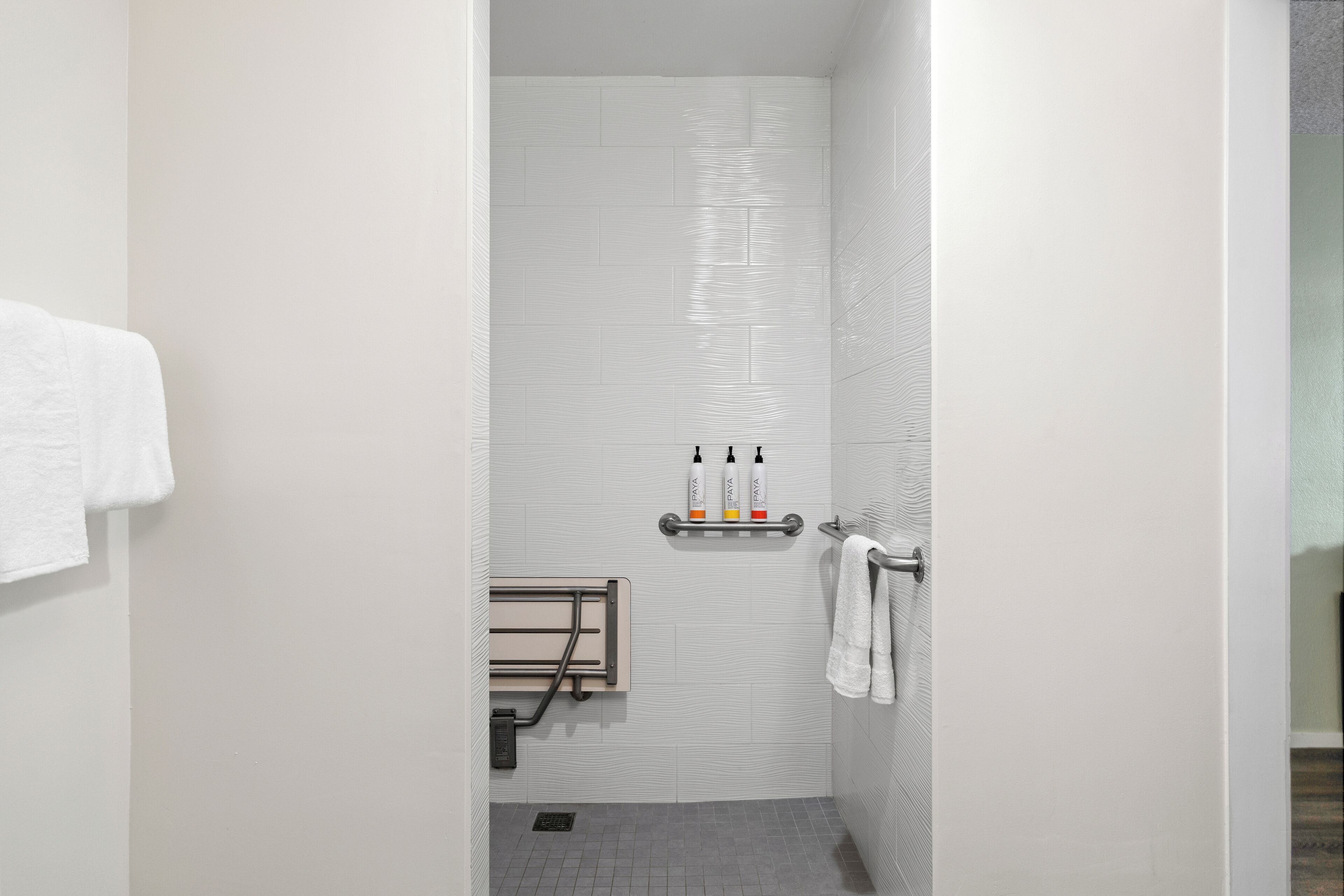 Un box de ducha estrecho, con azulejos blancos, un perchero y un estante con artículos de aseo.