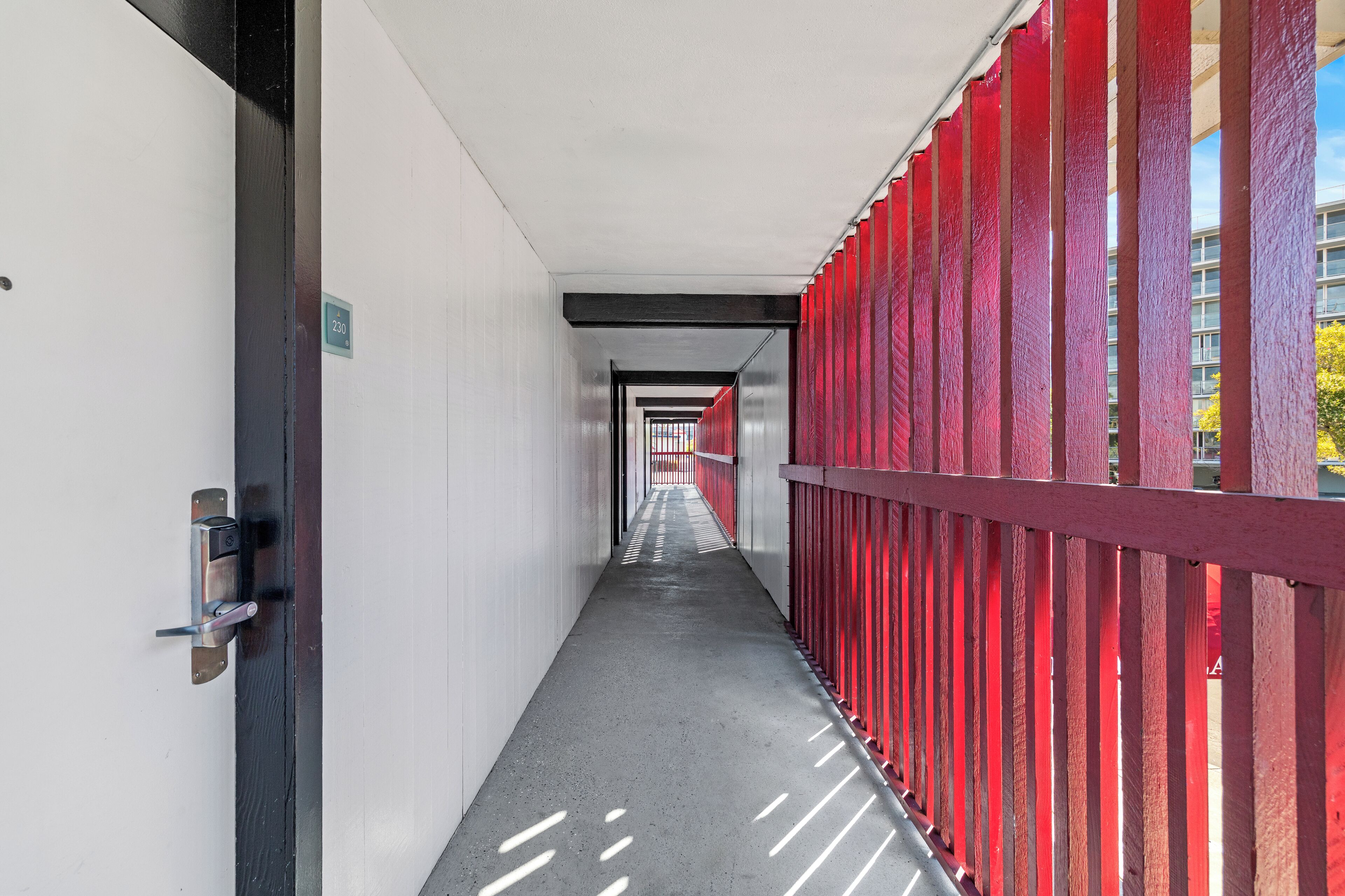 A corridor with a door on the left, a red railing on the right, and a building in the background.