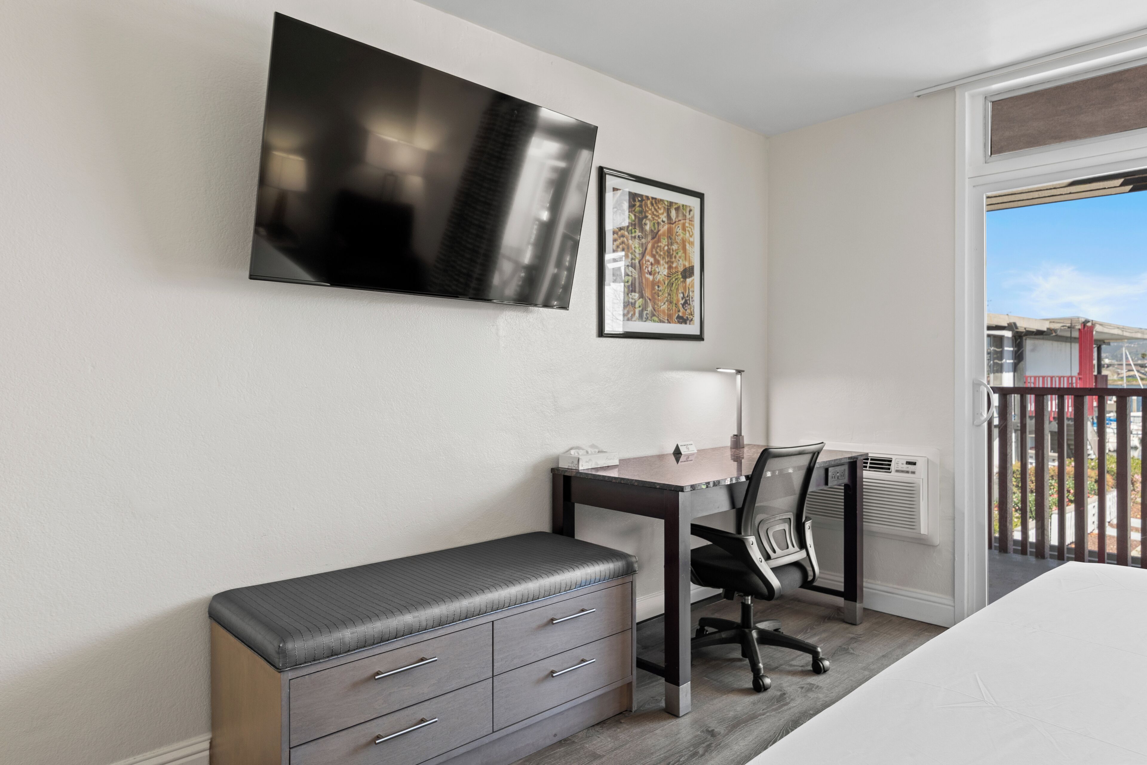 Una habitación de hotel moderna con una televisión de pantalla plana, un escritorio con silla, un banco con cajones y un ventanal con vistas al exterior.
