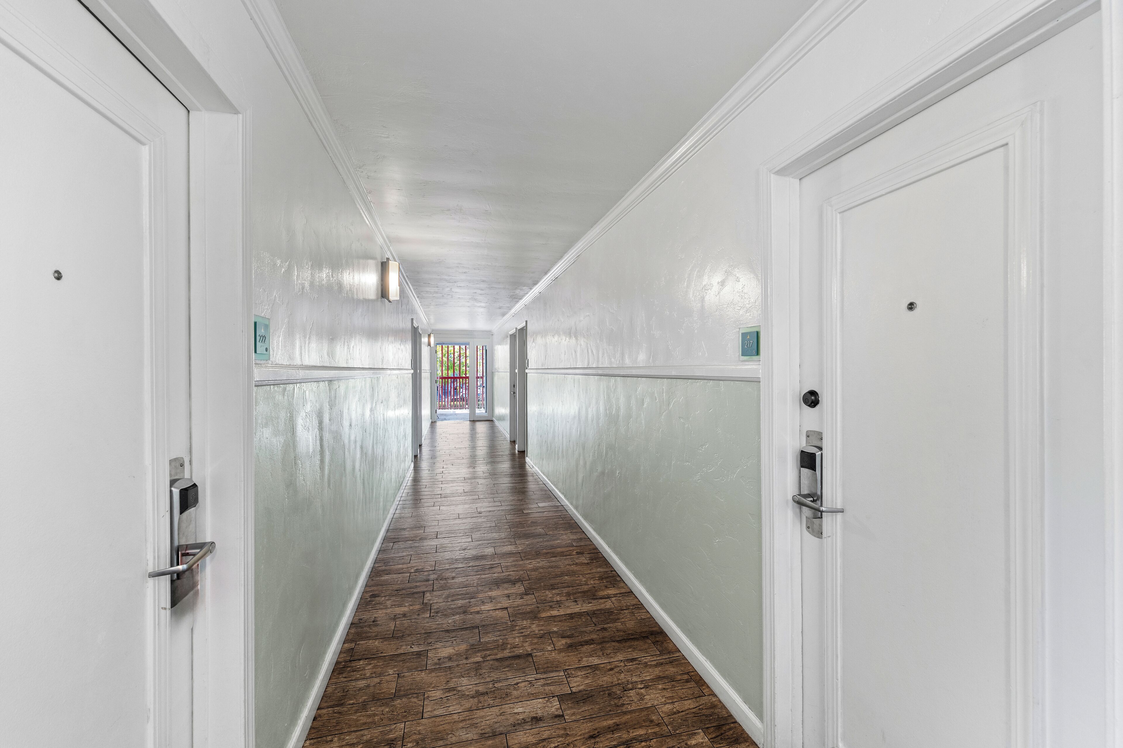 Un long couloir étroit aux murs blancs et au plancher en bois, qui mène à une pièce bien éclairée au bout.