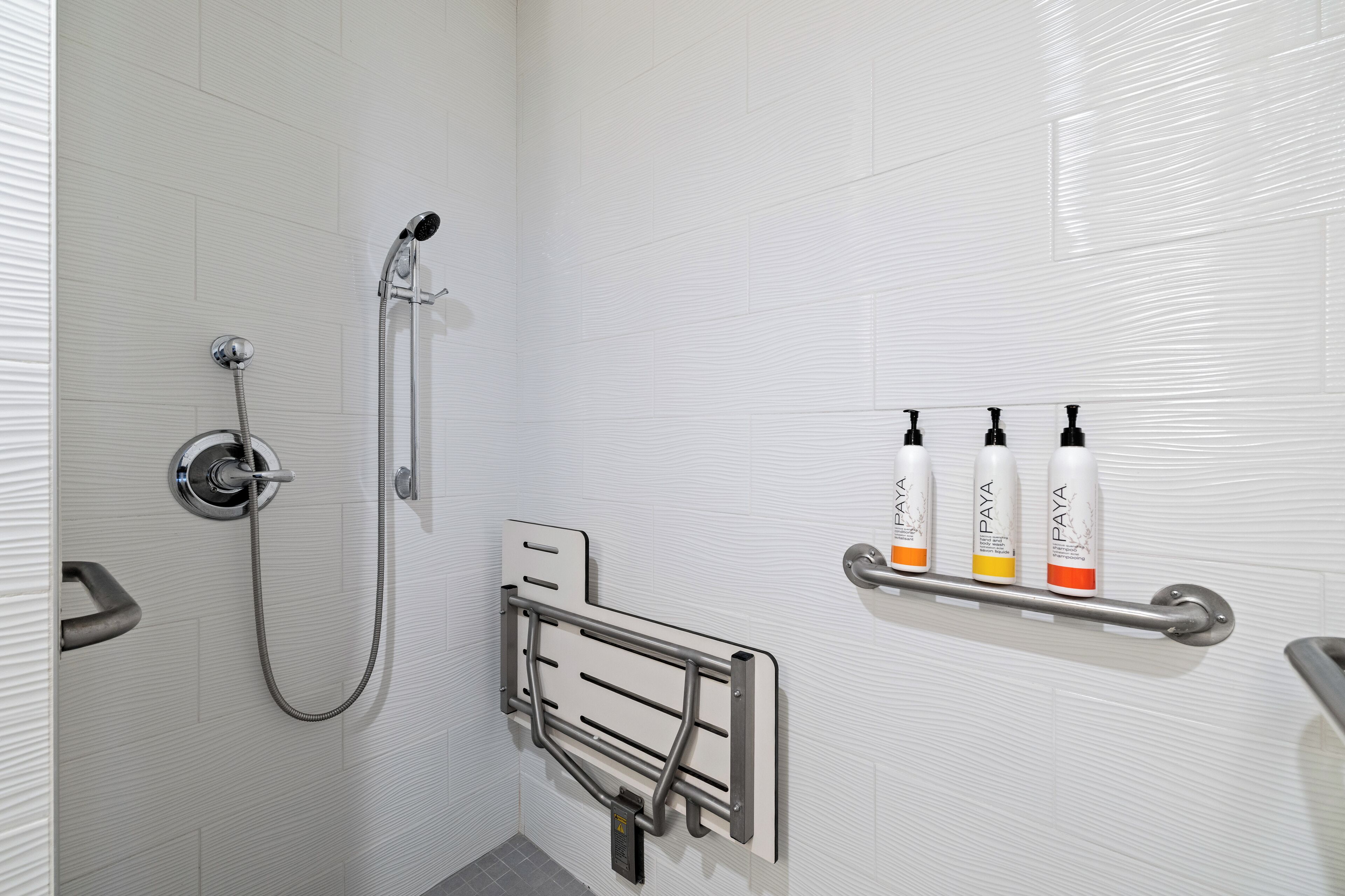 Une salle de bain avec une douche, une barre d’appui et trois bouteilles de shampoing sur une étagère.