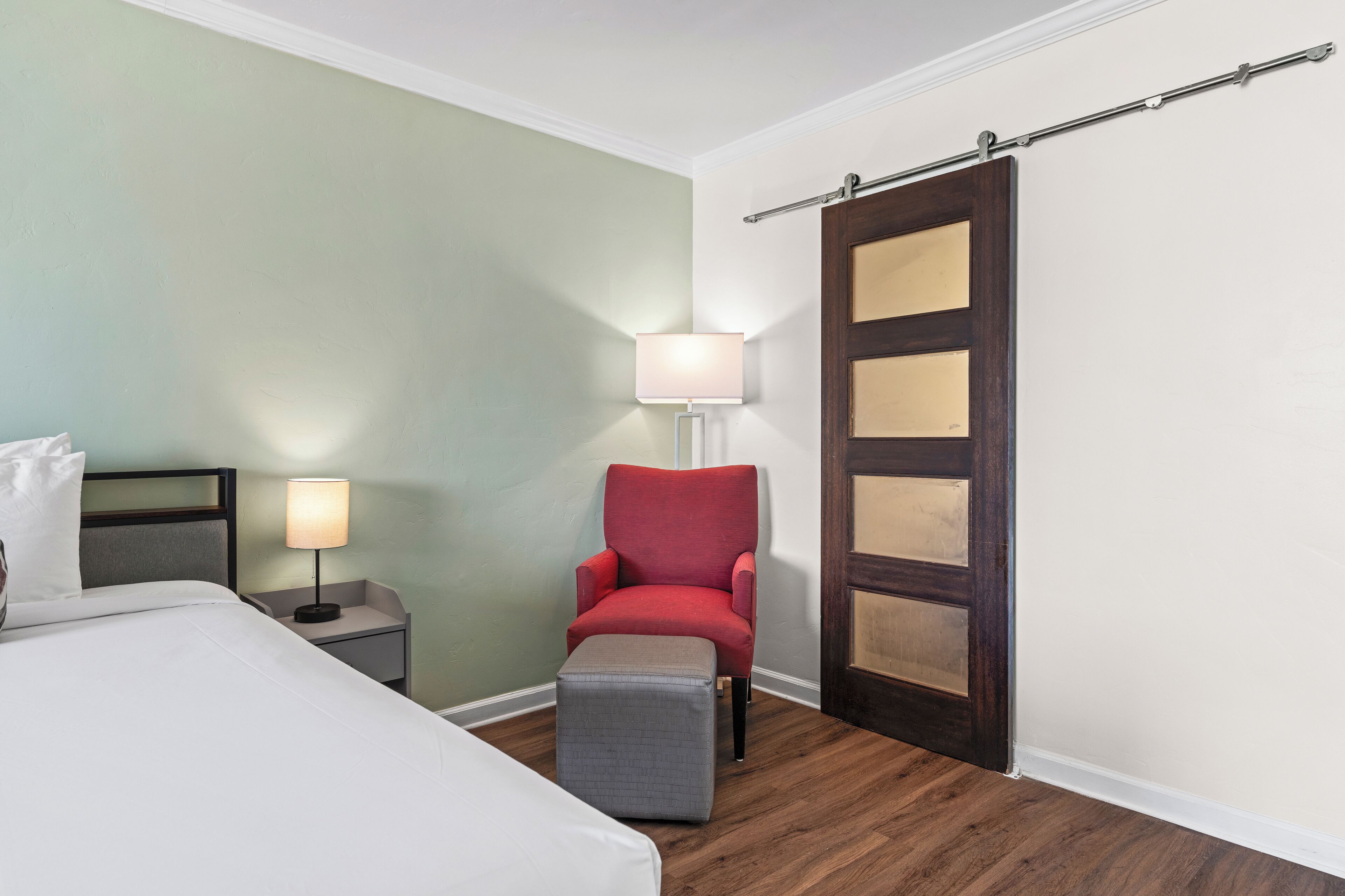 A hotel room with a bed, a red armchair, a small ottoman, a nightstand with a lamp, and a wooden door.