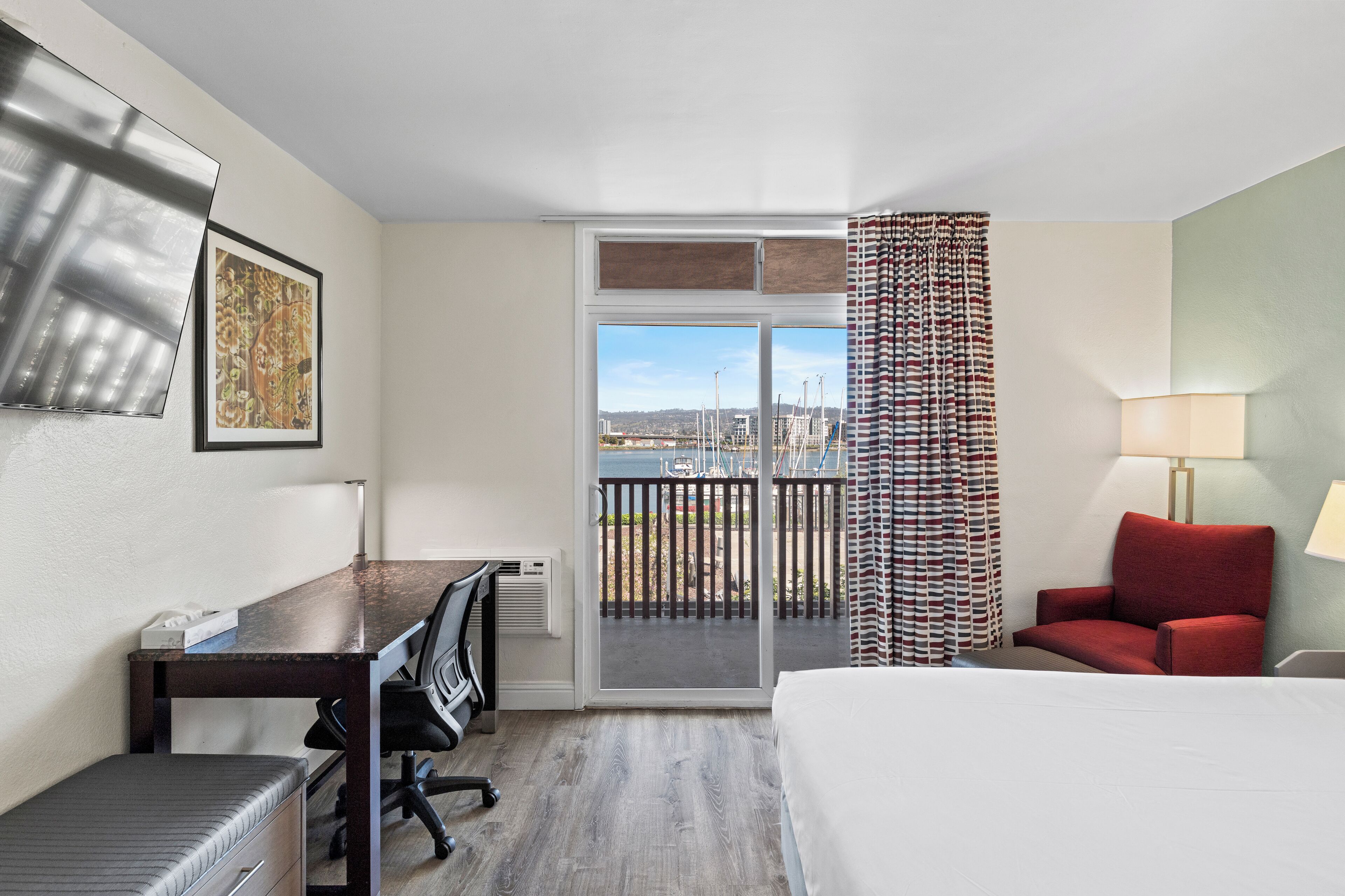 A hotel room with a bed, desk, chairs, a view through a sliding door, and a red chair.