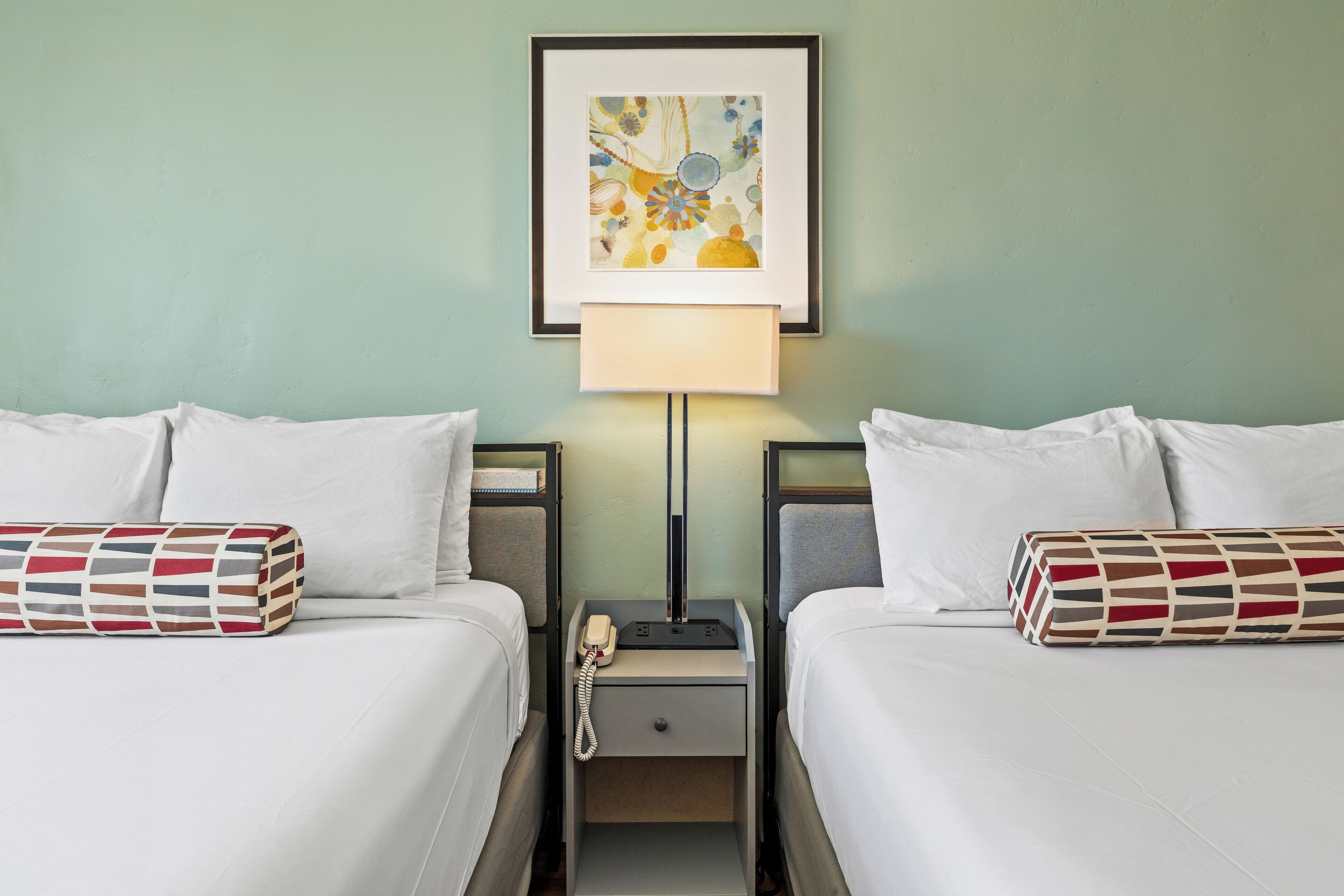 Hotellihuone, jossa on kaksi sänkyä, yöpöytä lampun kanssa ja seinällä oleva kehystetty taideteos.