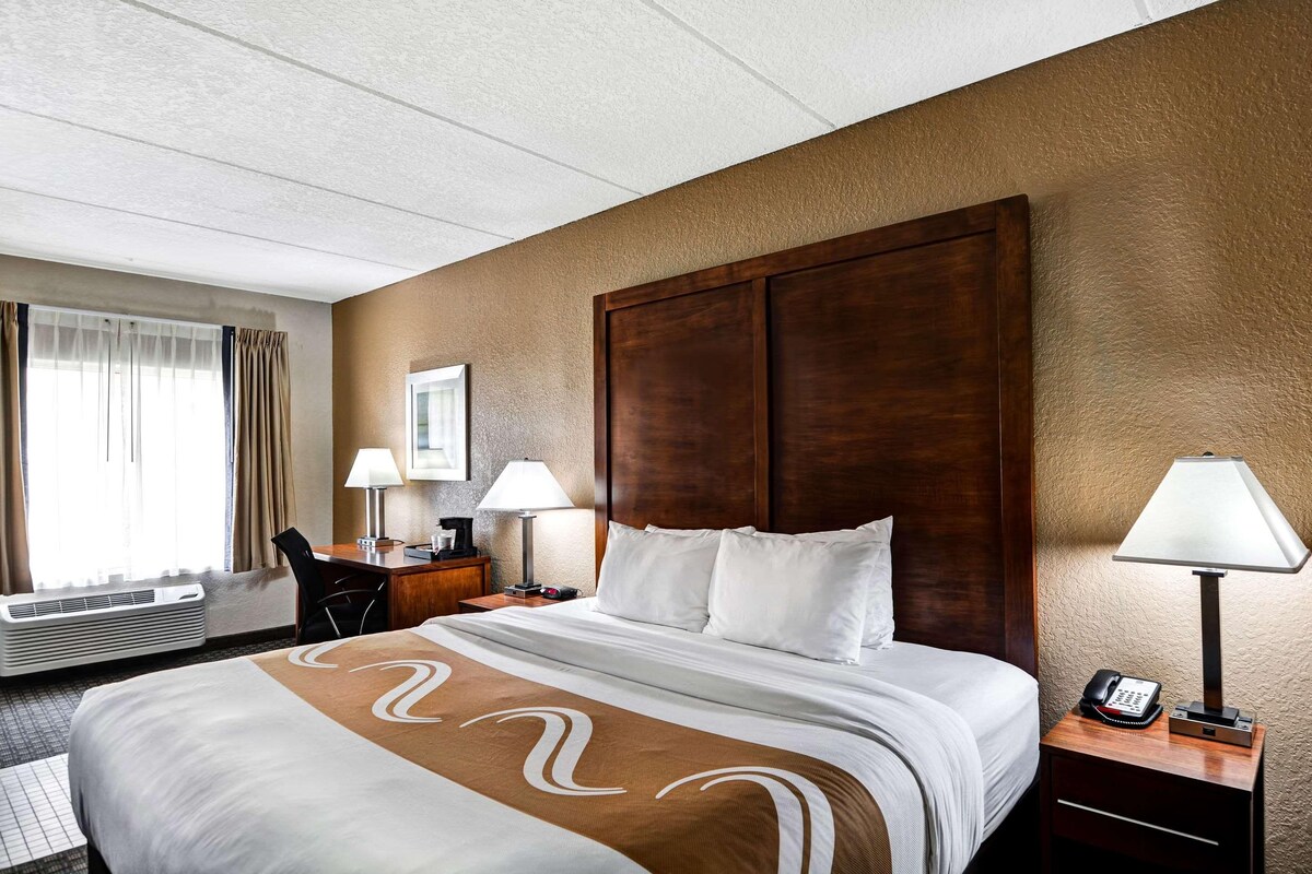 A hotel room with a large bed, two bedside lamps, a desk with a chair, a telephone, and a window with curtains.