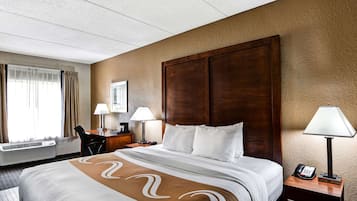 A hotel room with a large bed, a desk with a chair, a desk lamp, a phone, and a window with curtains.