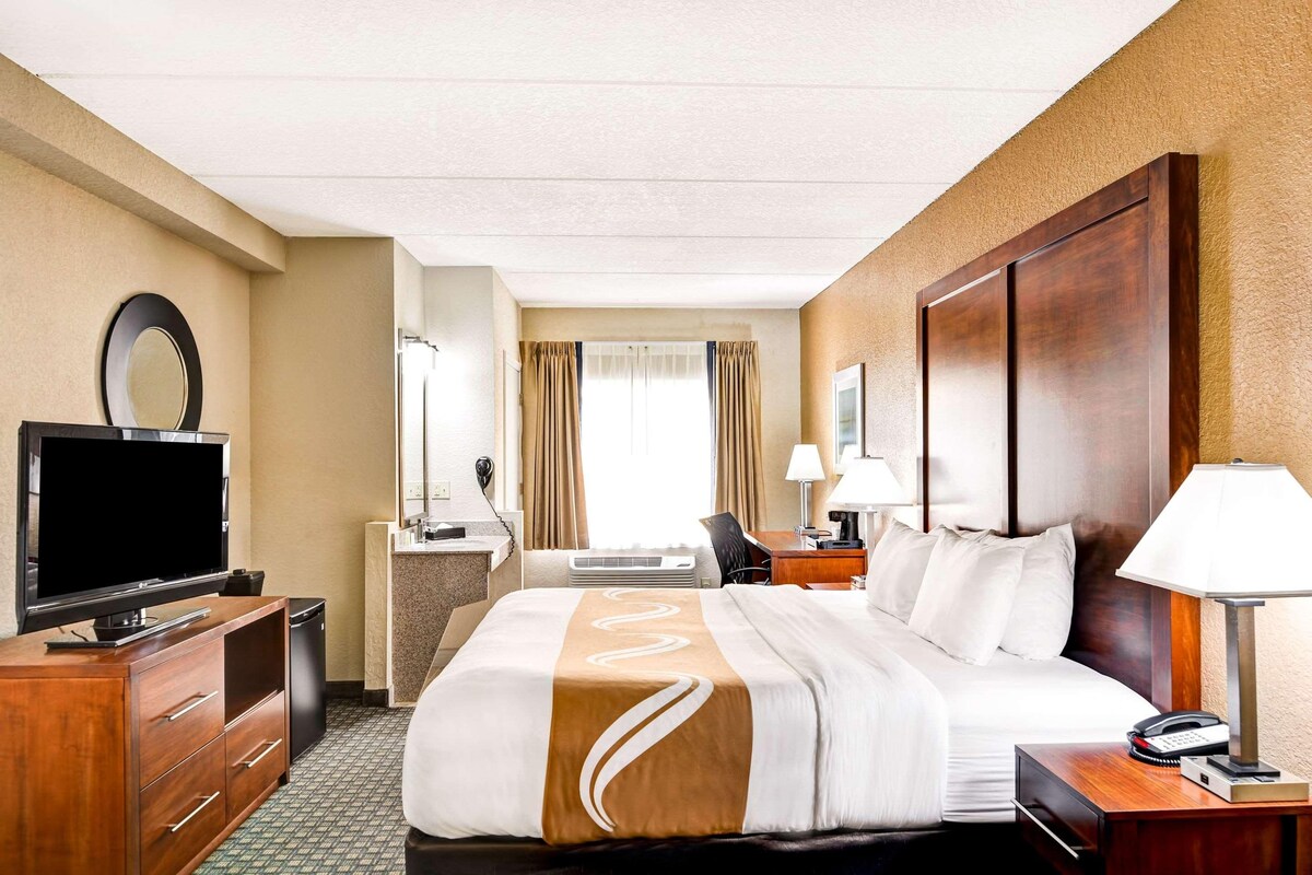 A hotel room with a bed, a TV, a desk with a lamp, and a bathroom in the background.