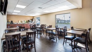 A restaurant with multiple tables and chairs, a kitchen area, and a large window providing a view of the outside. - Quality Inn & Suites Heritage Park (Kissimmee)