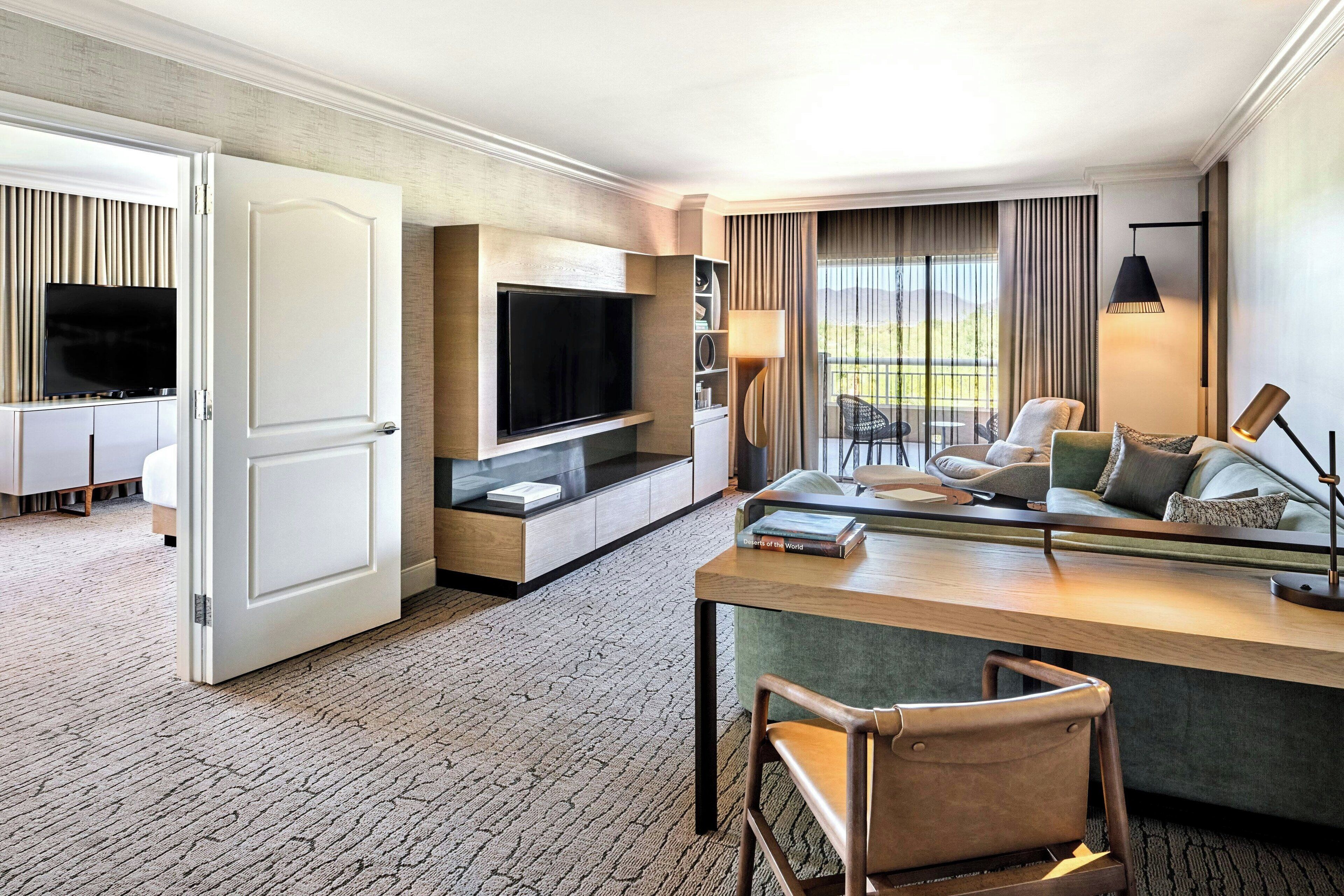 A modern hotel room with a large flat-screen TV, a comfortable seating area with a sofa and chairs, a small desk with a lamp, and a view of the outdoors through the window.