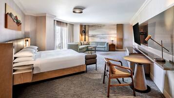 A hotel room with a large bed, a seating area with a table and chairs, a flat-screen TV mounted on the wall, and a large window with curtains.
