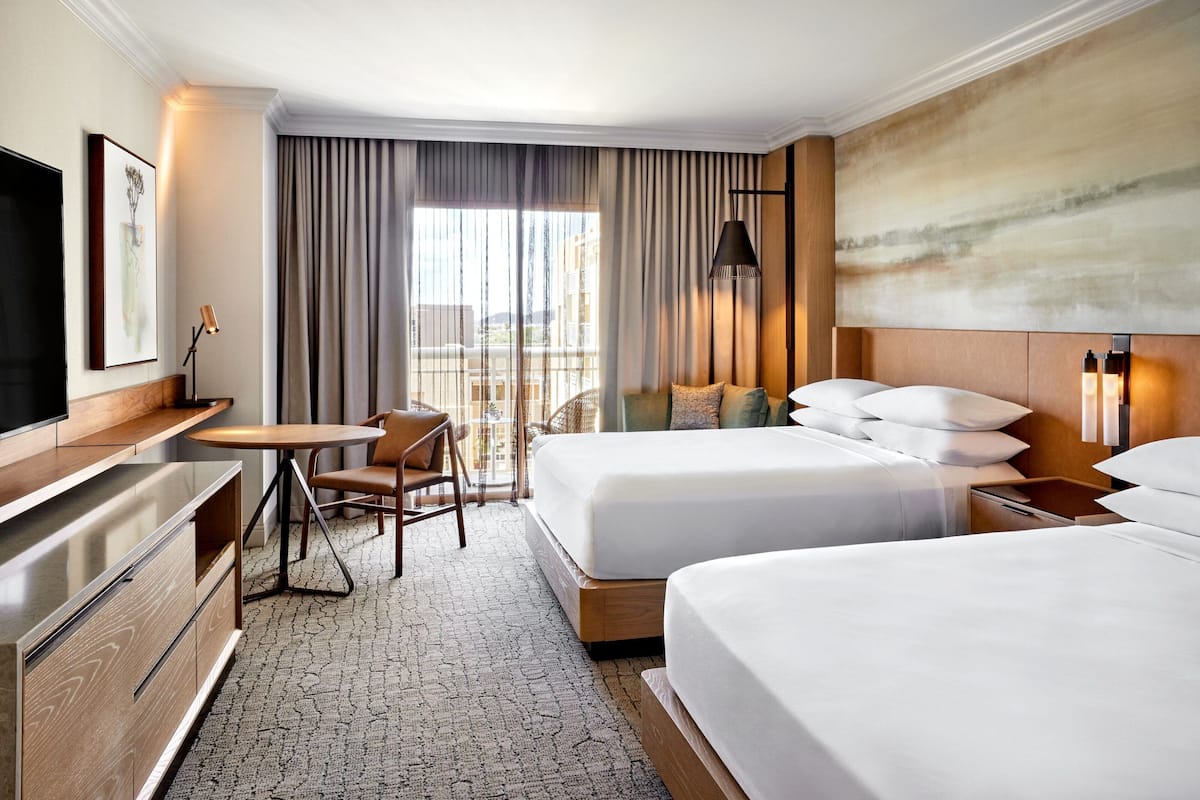 A hotel room with two beds, a desk, a chair, a TV, and a large window with curtains.