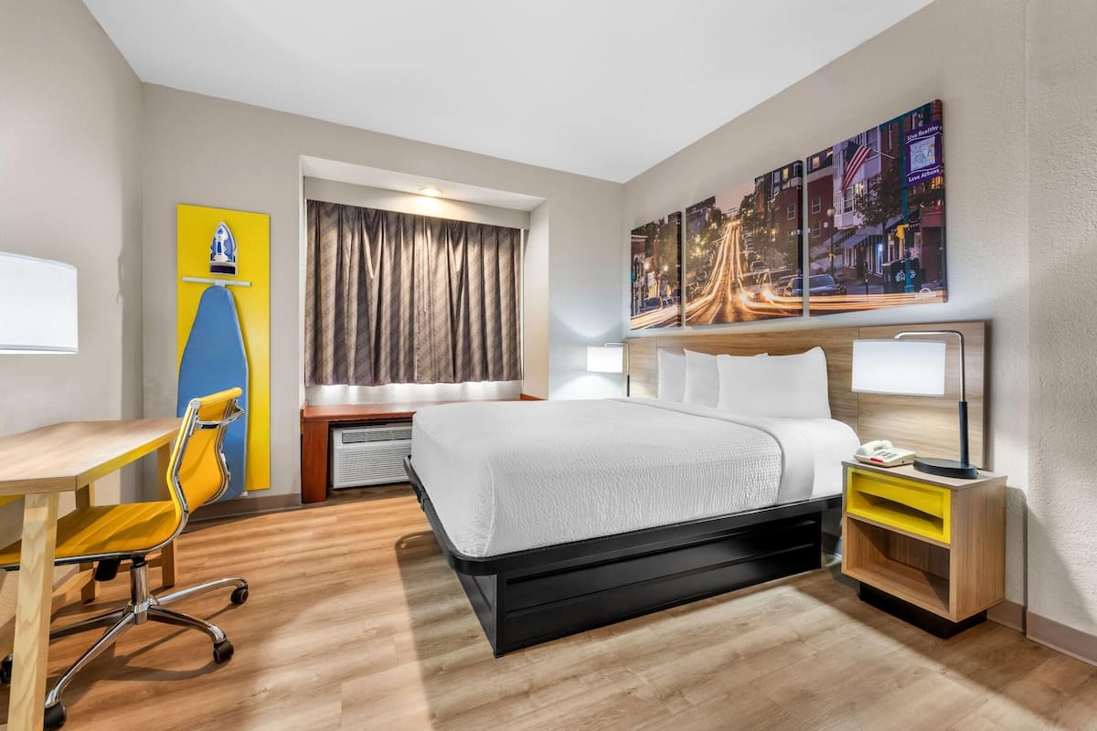 A hotel room with a bed, desk, chair, and artwork on the wall.