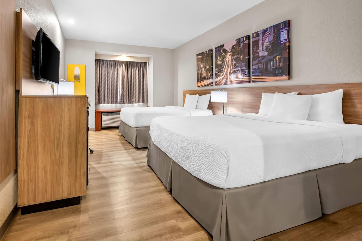 A hotel room with two beds, a TV, a desk, a window with curtains, and a cityscape painting on the wall.