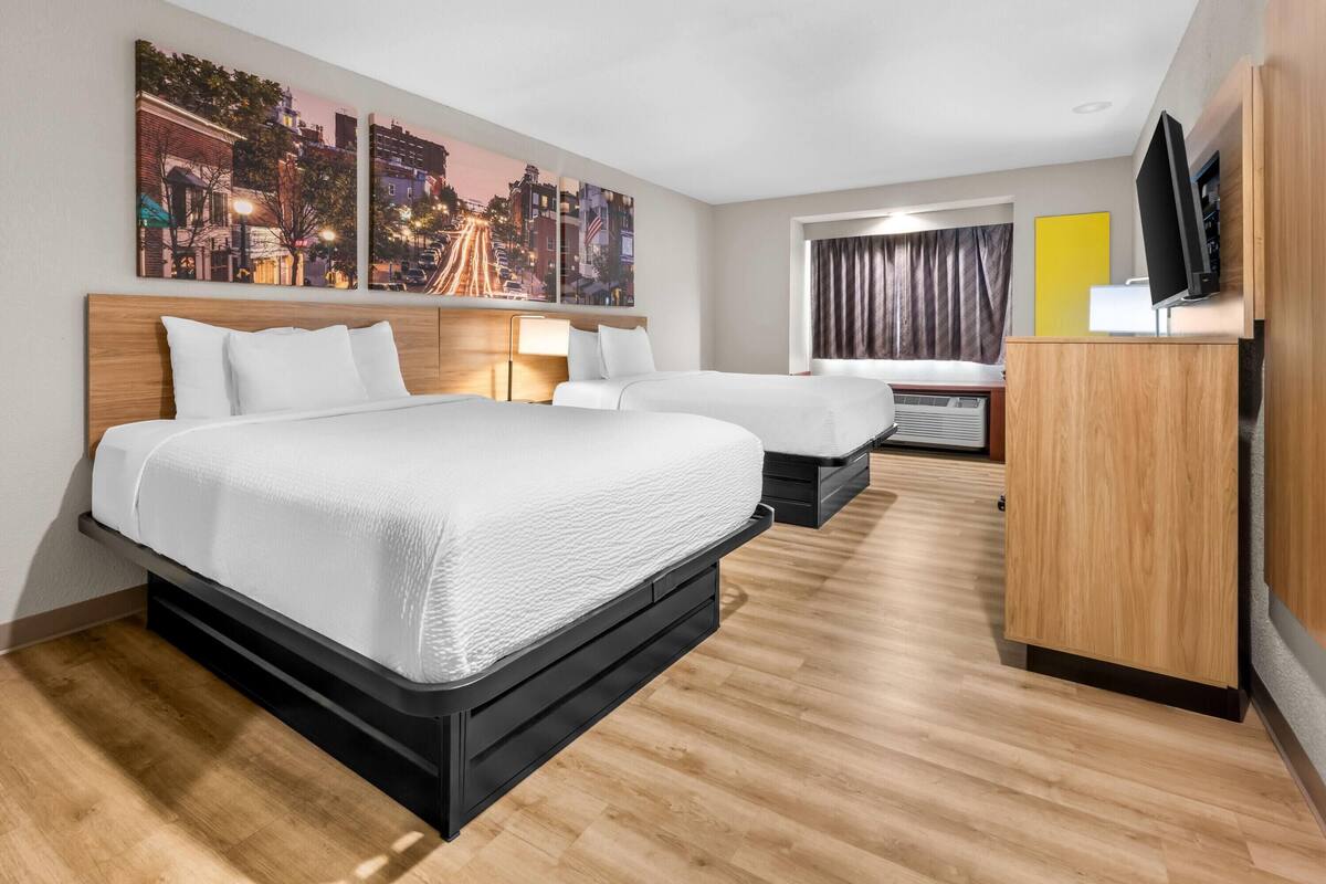 A hotel room with two beds, a TV, a desk, and artwork on the wall.