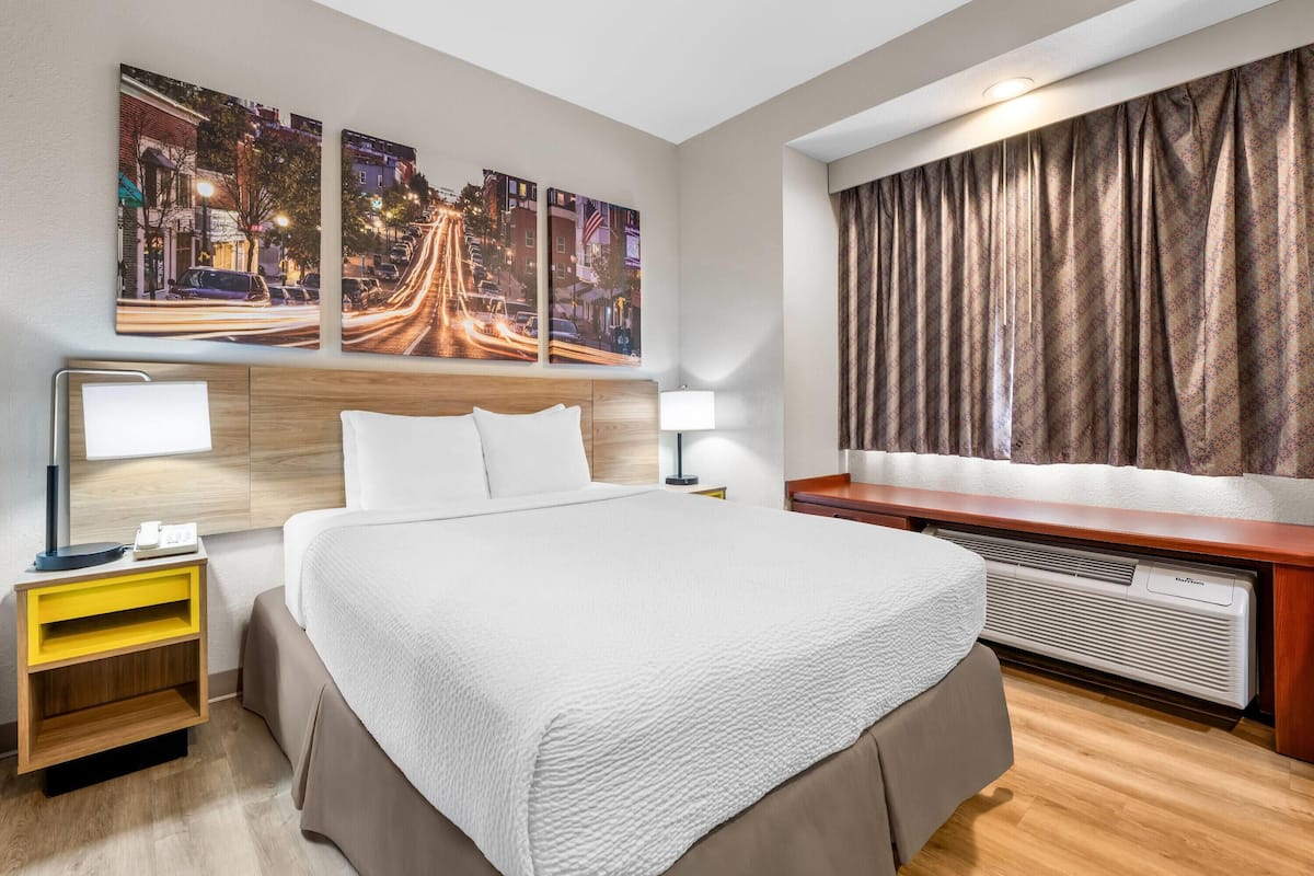 A hotel room with a large bed, bedside tables, a heater, and cityscape artwork on the wall.