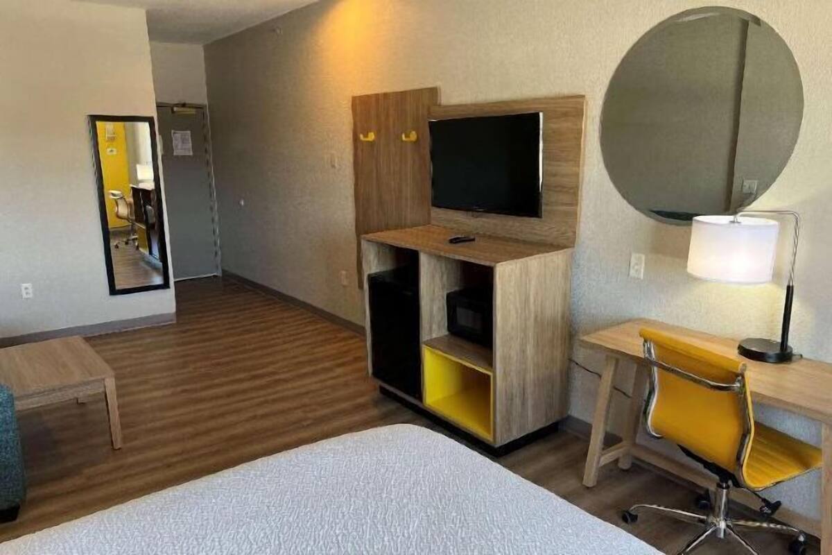 A hotel room with a bed, a desk with a chair, a TV, a mirror, and a lamp.