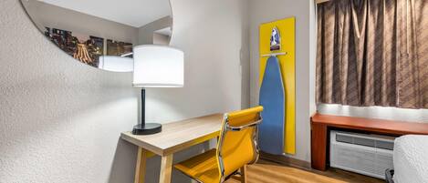 A hotel room with a wooden desk, a yellow chair, a mirror, a lamp, a bed, a heater, and a view of buildings through a window.
