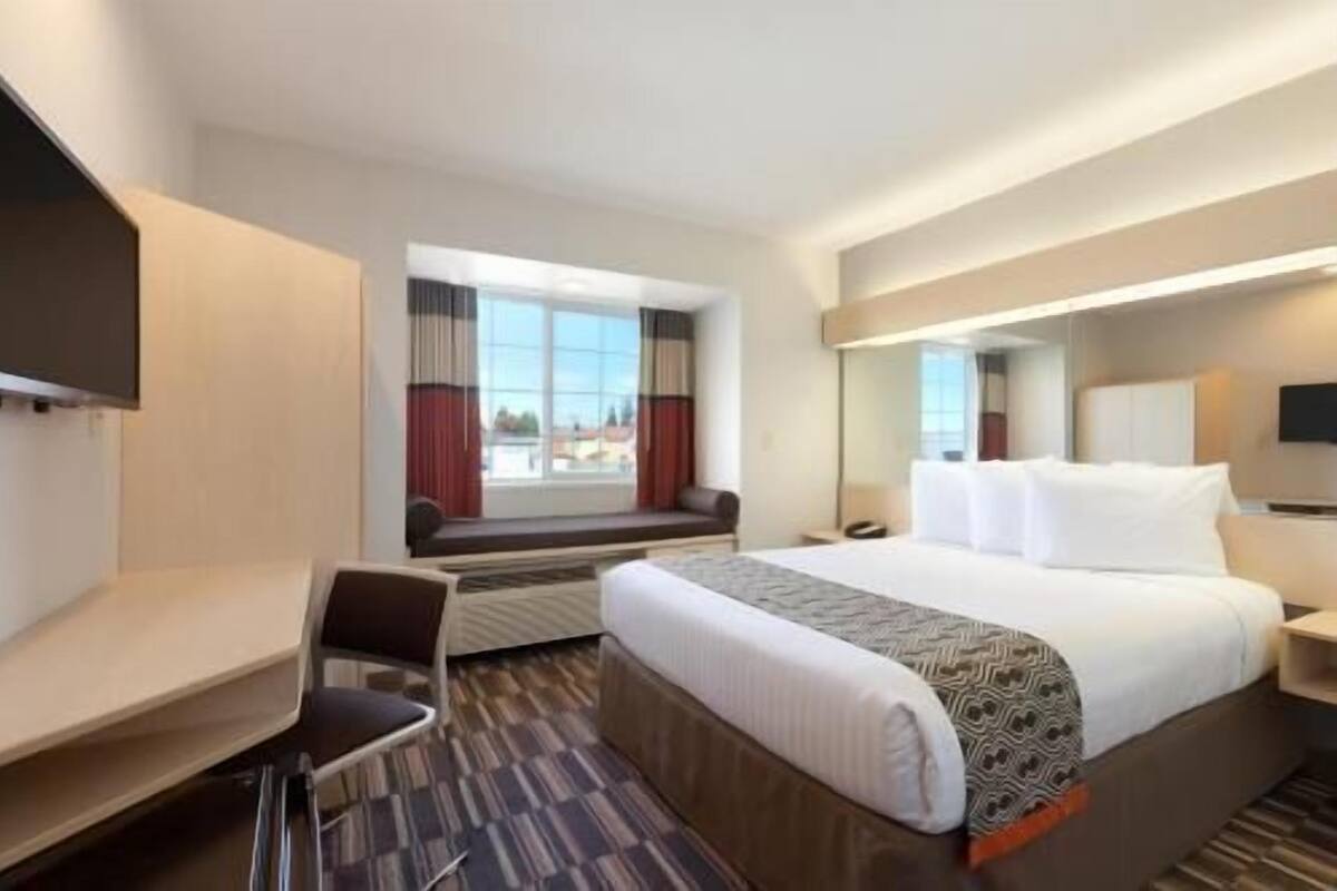Habitación de hotel con cama, escritorio, silla, televisión y ventana con cortinas.