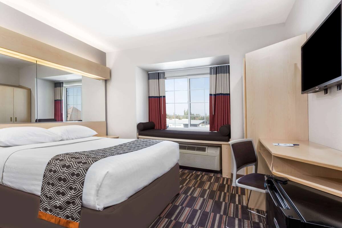 Una habitación de hotel con cama, escritorio, silla, televisión y ventana con cortinas.