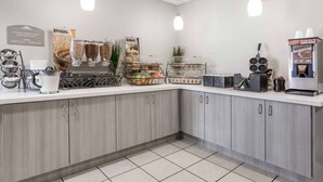 A breakfast bar with a complimentary breakfast sign, a variety of grains, a bakery display, and a coffee machine.