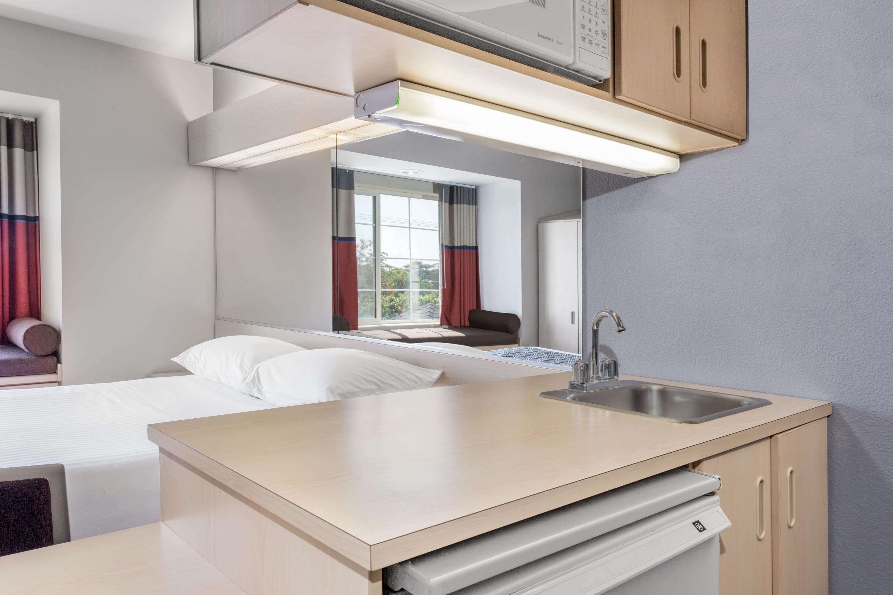Ein kleines Hotelzimmer mit Bett, einer kleinen Küchenzeile, einem Fenster mit Vorhängen und Blick ins Freie.