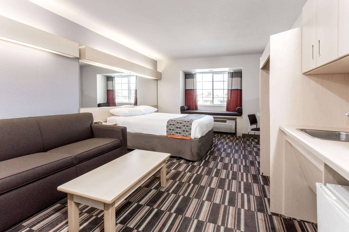 Una habitación de hotel compacta con sofá, cama individual, escritorio pequeño, kitchenette y un ventanal grande con cortinas.