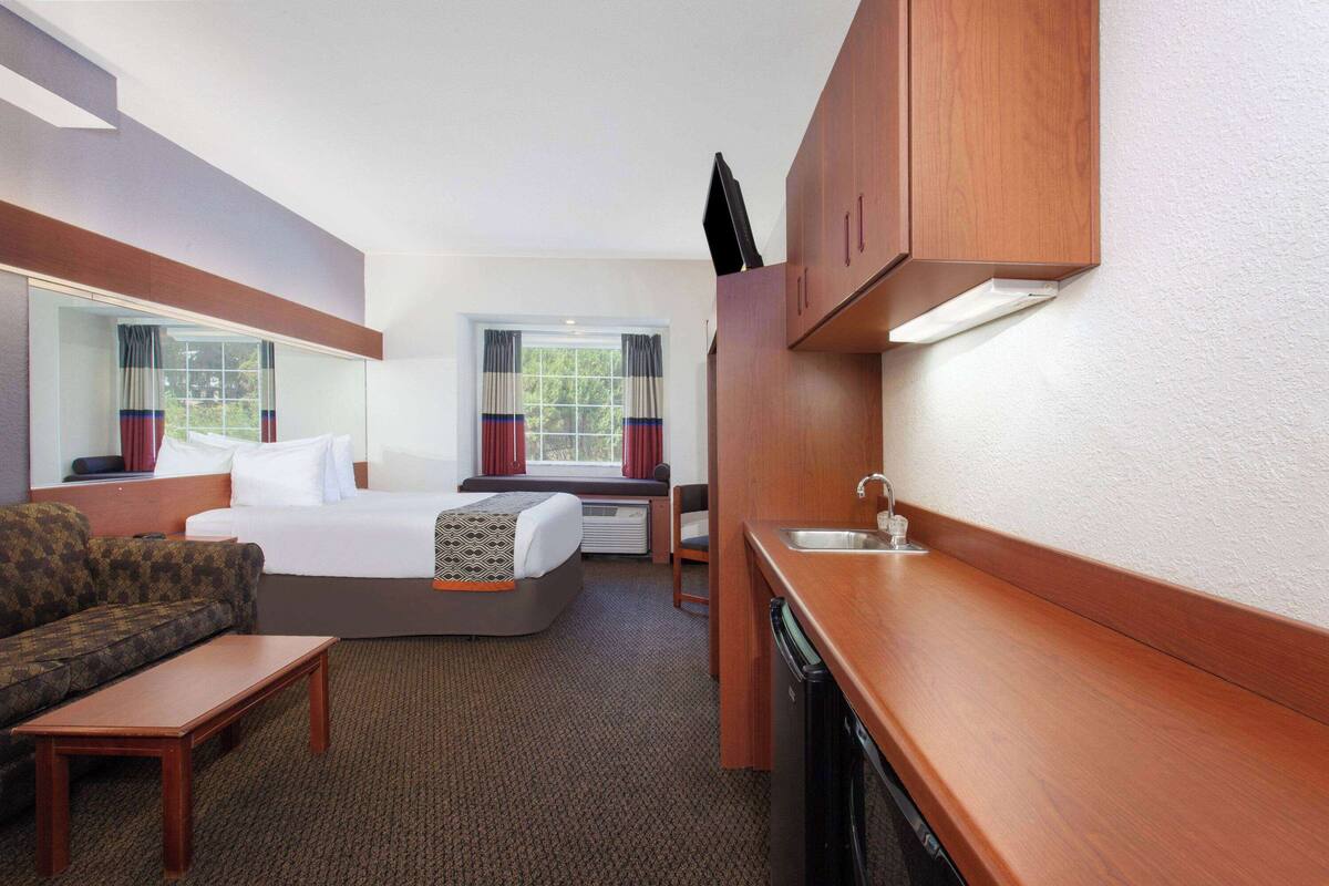 A compact hotel room with a bed, a sofa, a small table, a kitchenette, and a window with curtains.