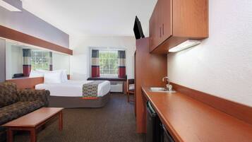 Quarto de hotel pequeno com cama, sofá, mesa, cozinha compacta e janela com cortinas.
