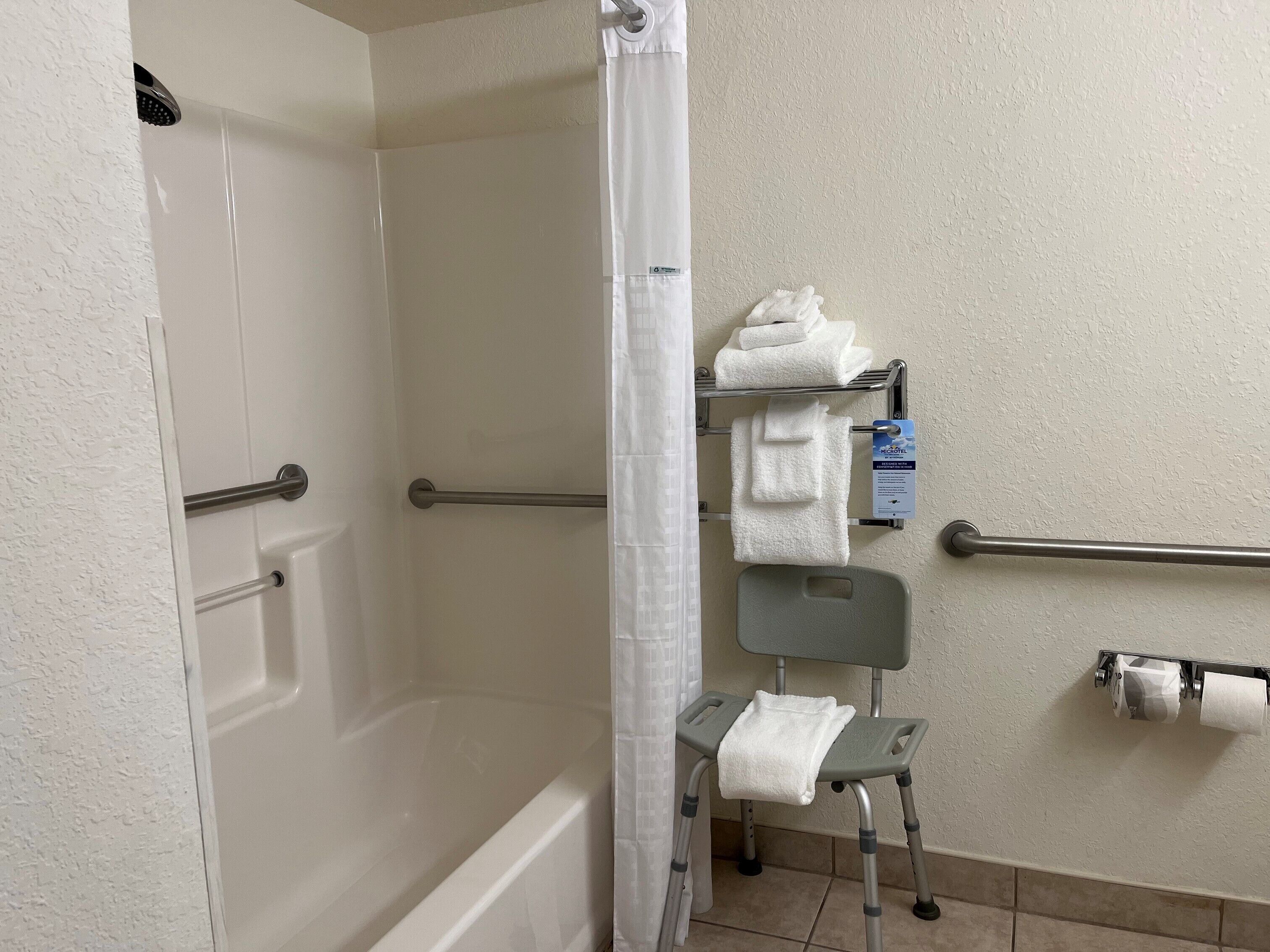 Un baño con ducha, barras de apoyo, un asiento accesible para silla de ruedas y un perchero para toallas.