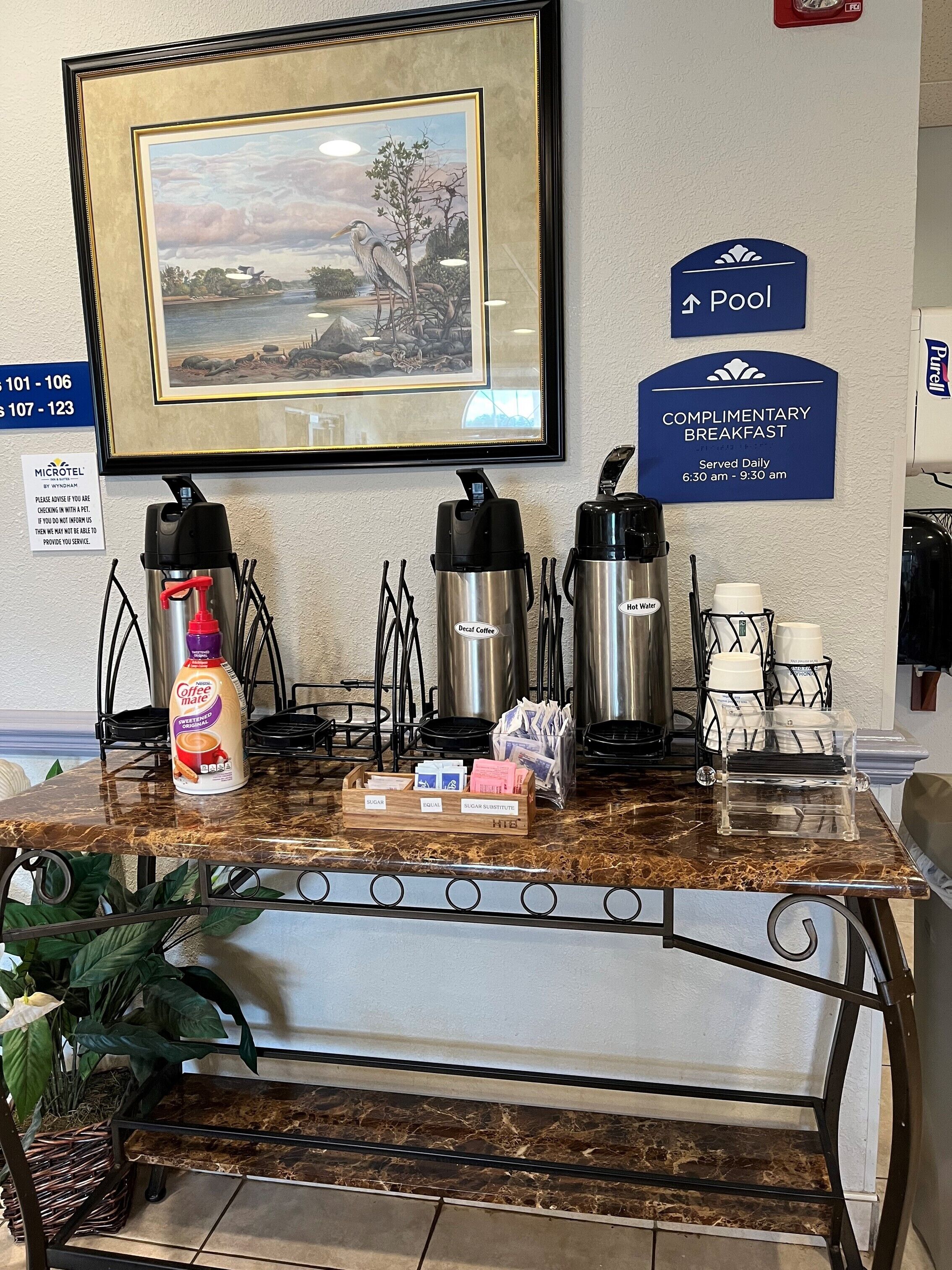 Zona de recepción de hotel con estación de café, un cuadro enmarco con paisaje, un cartel anunciando desayuno de cortesía y una señal que indica la ubicación de la piscina.