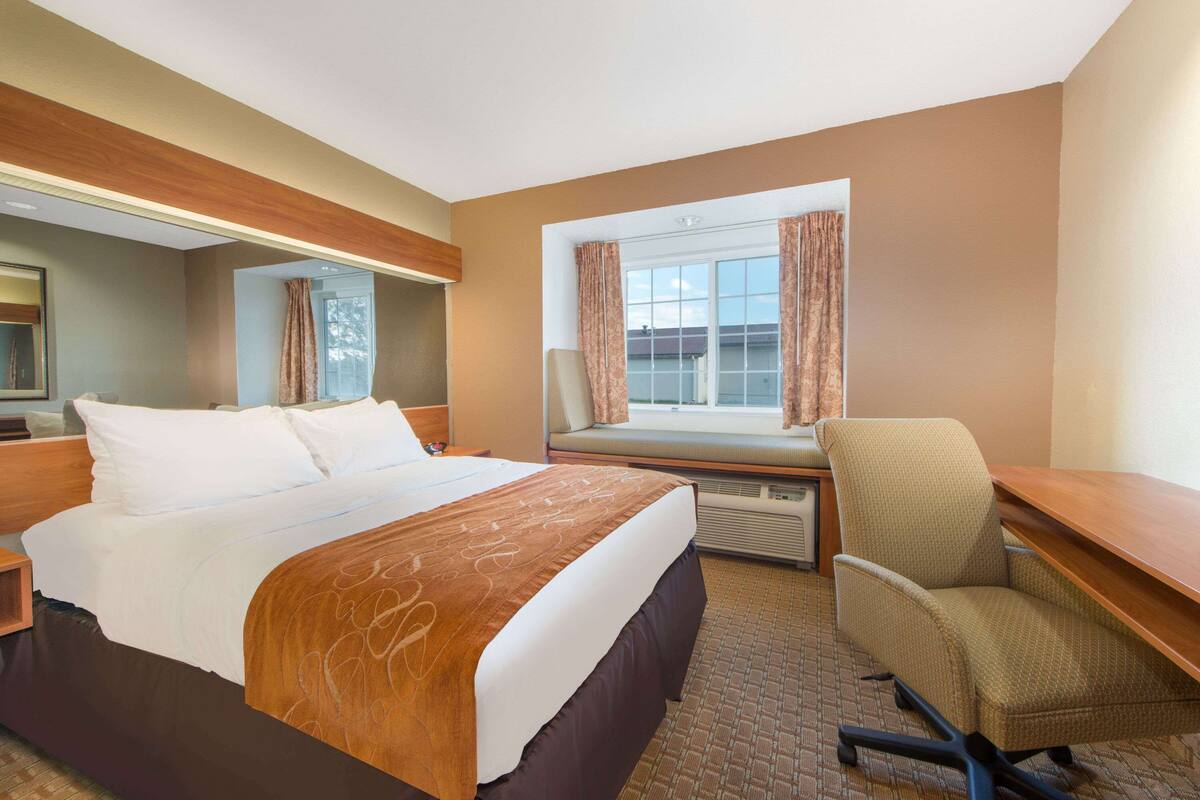 A hotel room with a large bed, a desk, a chair, a window with curtains, and a mirror.