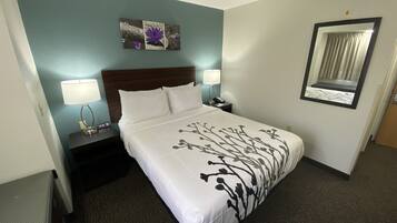 Una camera d'hotel con un letto, due abat-jour, un comodino, uno specchio a parete e un quadro decorativo sopra il letto.