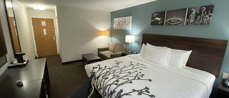 A hotel room with a bed, a desk, a chair, a lamp, and a wall with artwork.