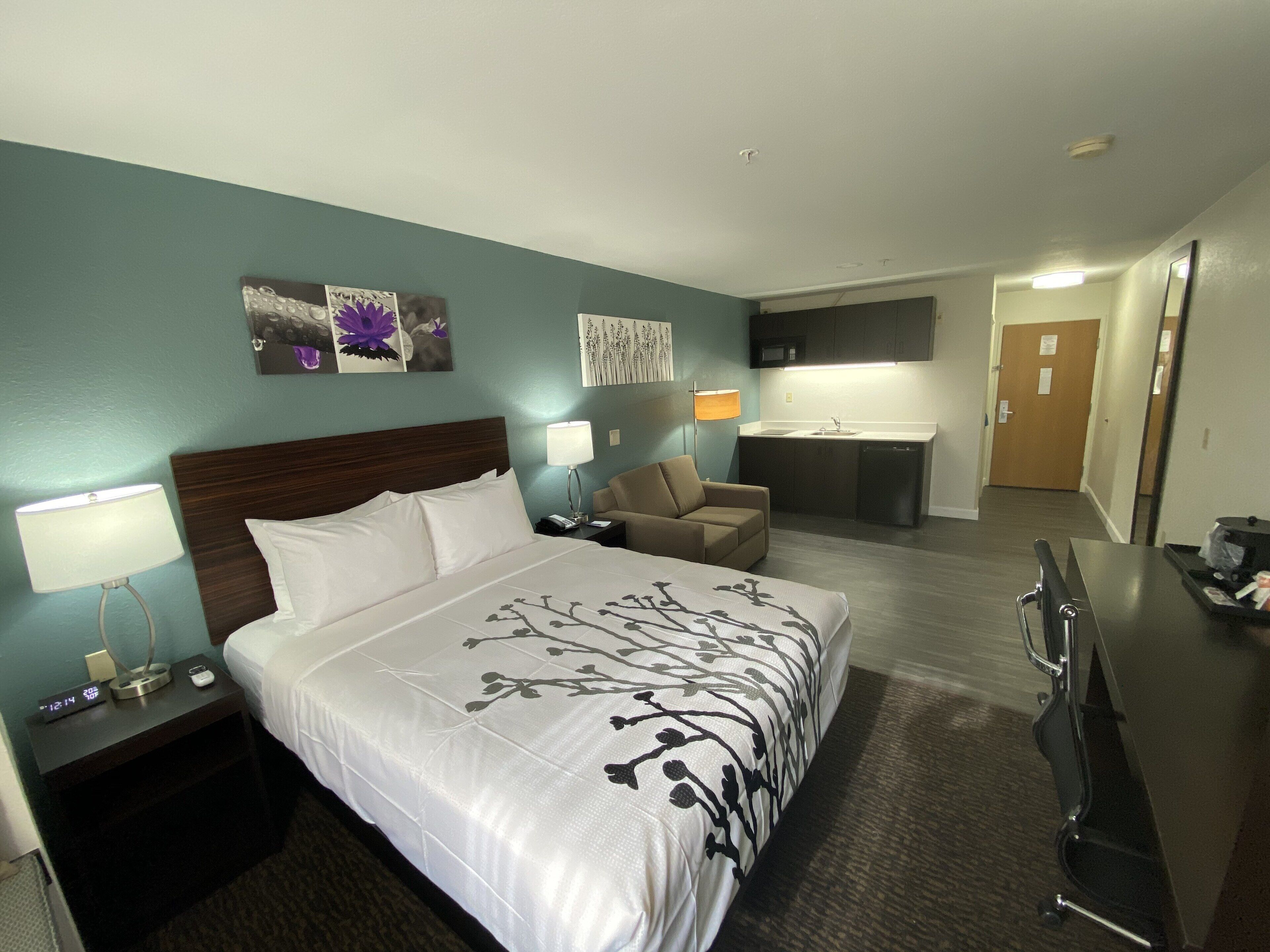 A hotel room with a bed, a desk, a chair, a nightstand, and a kitchenette.