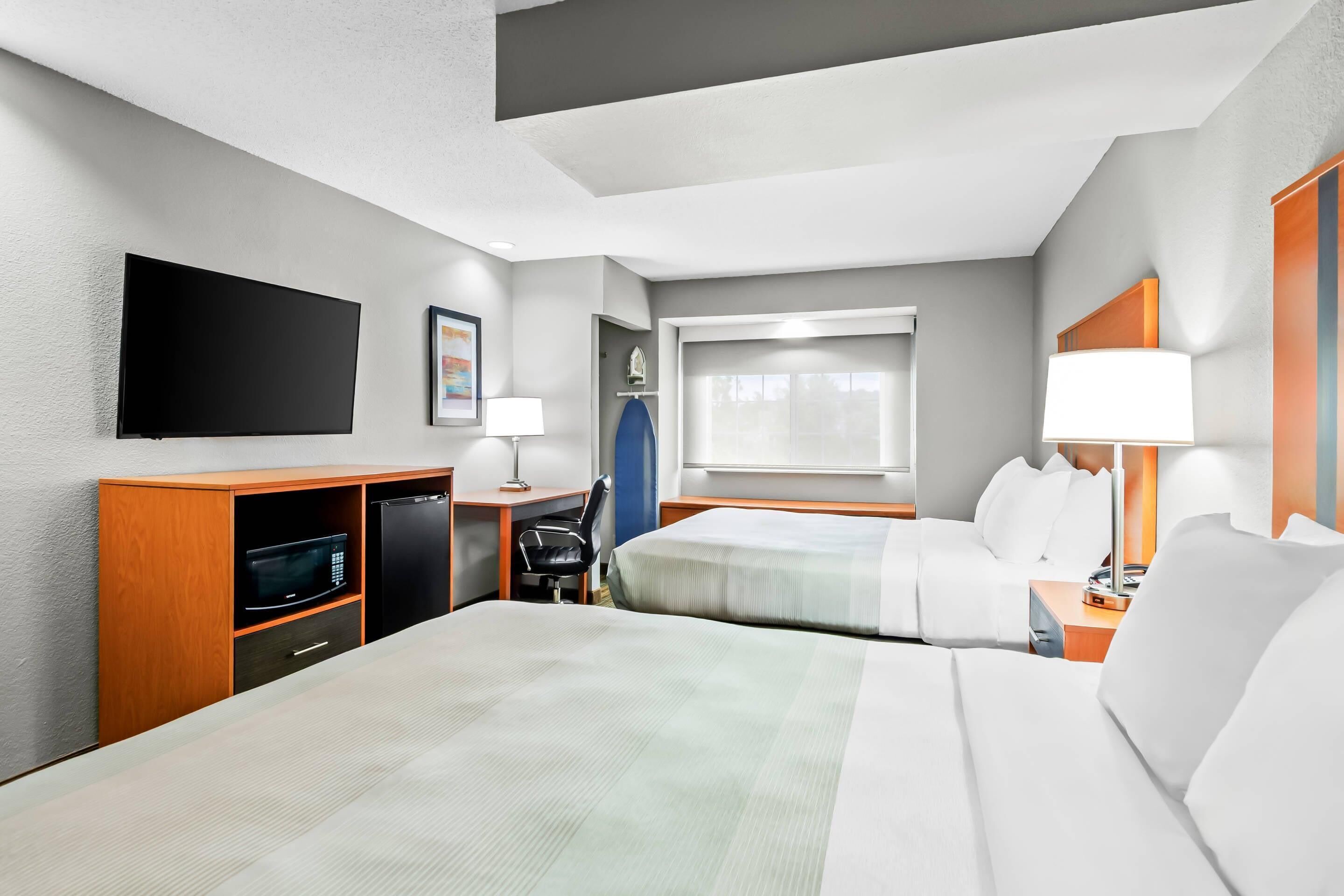 Habitación de hotel con dos camas, un televisor de pantalla plana, un escritorio con silla, un microondas y una ventana con persianas.