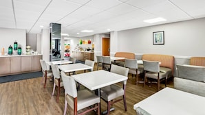 Un espace repas moderne avec des tables et des chaises blanches, une cuisine en arrière-plan et un tableau encadré au mur.