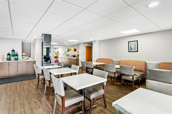 Un espace repas moderne avec des tables et des chaises blanches, une cuisine en arrière-plan et un tableau encadré au mur.