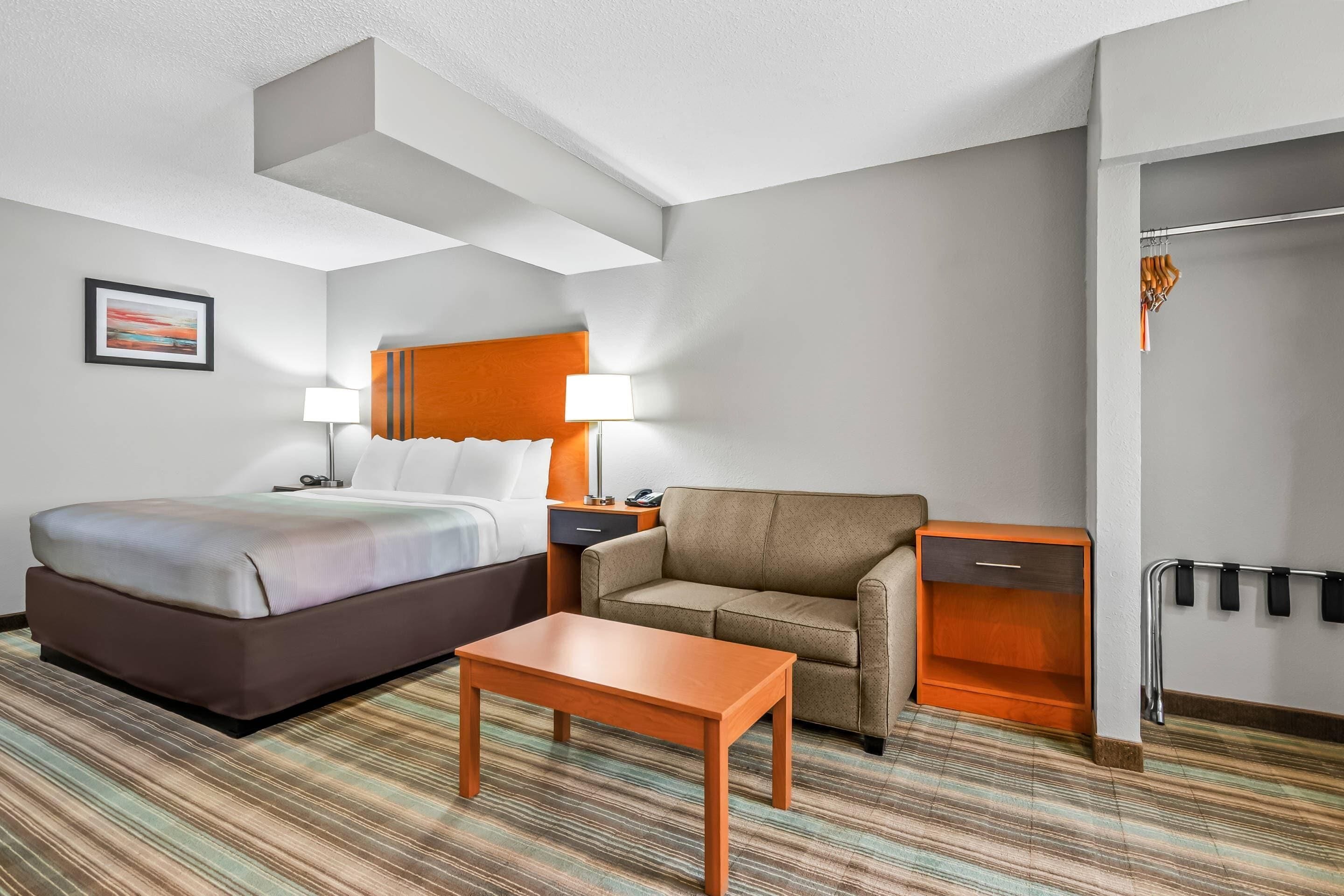 Ein Hotelzimmer mit Bett, Sofa, Tisch, Nachttisch, Lampe, Kleiderhaken und einem gerahmten Bild an der Wand.