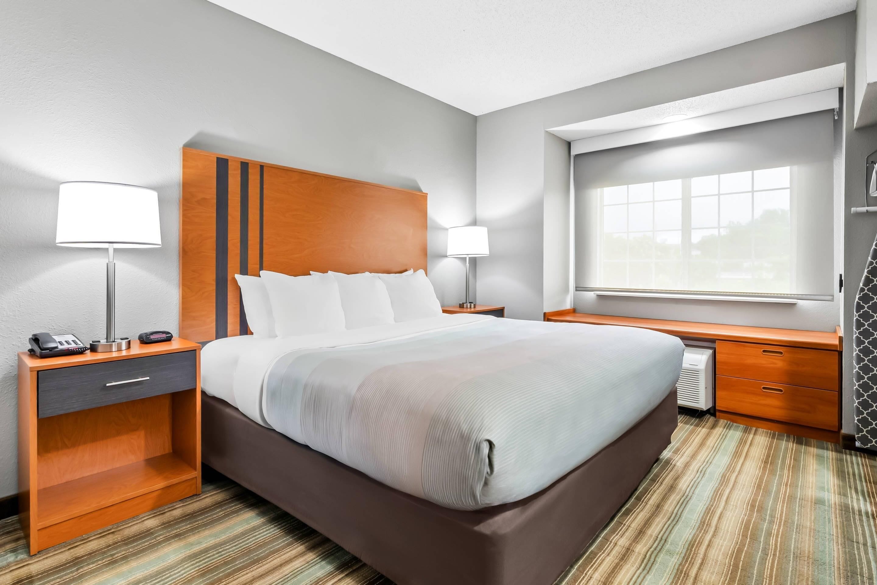 A hotel room with a large bed, two bedside tables with lamps, a window with a view, and a built-in desk with drawers.