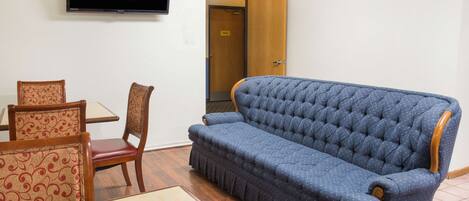 Une pièce avec un canapé bleu, des chaises en bois, une petite table et un téléviseur fixé au mur.
